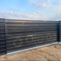 KC-SPAW - Ciemnoszara brama przesuwna z poziomymi panelami, zamontowana na betonowej podstawie w piaszczystym terenie, z widocznymi prowadnicami i fragmentem ogrodzenia.