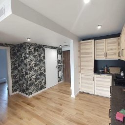 Konstantin Antonenko - Wykończone wnętrze mieszkania: drewniana podłoga, ściana z tapetą w roślinny wzór, jasne drewniane szafki kuchenne i białe drzwi. Widok na korytarz.