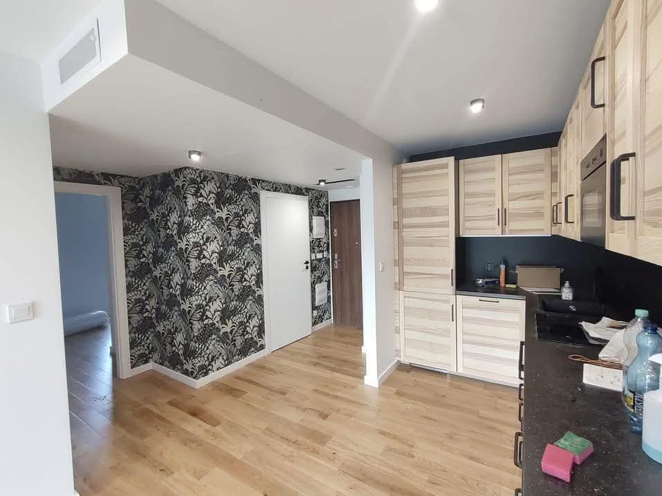 Wykończone wnętrze mieszkania: drewniana podłoga, ściana z tapetą w roślinny wzór, jasne drewniane szafki kuchenne i białe drzwi. Widok na korytarz.