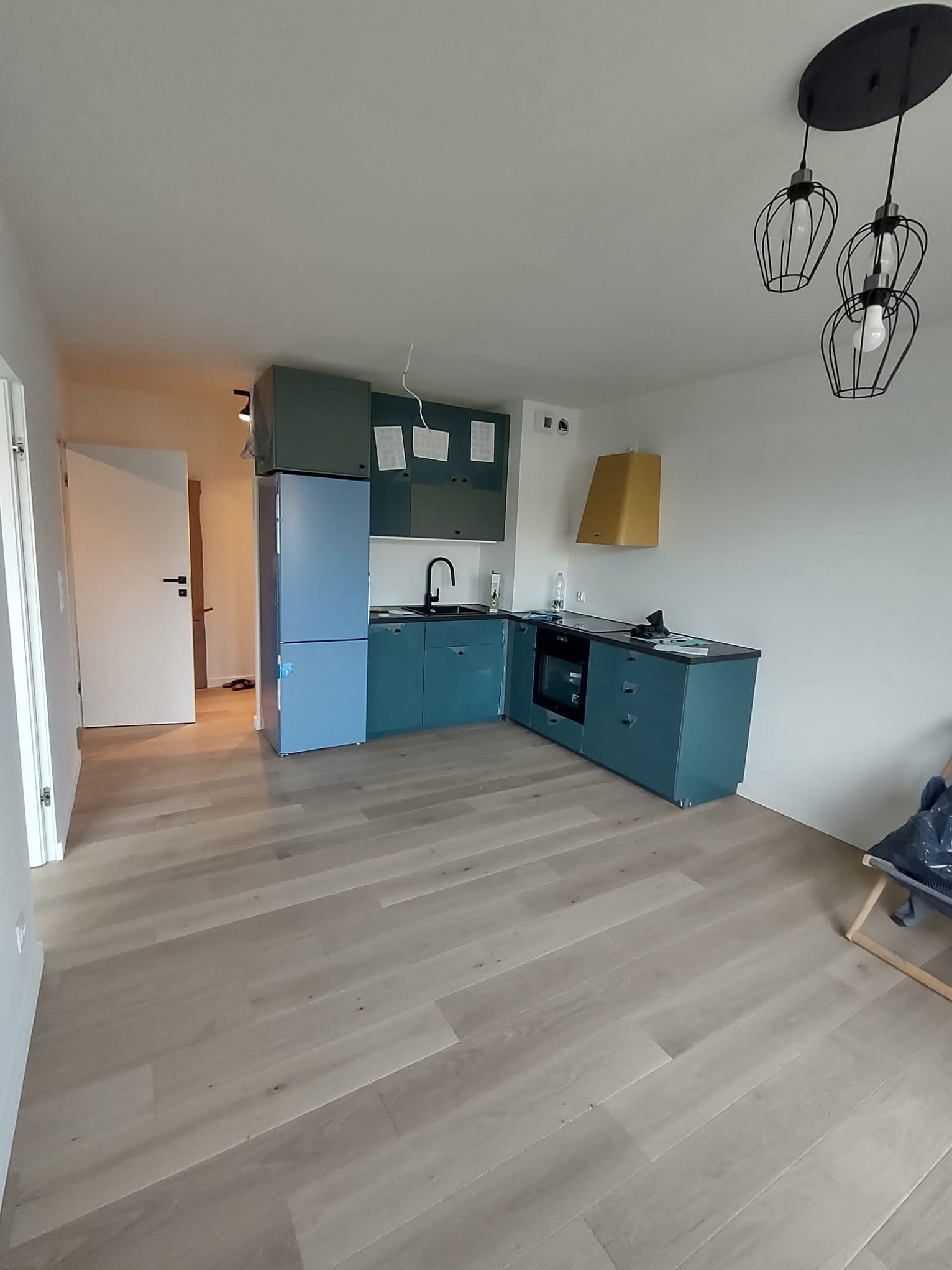 Niebieska zabudowa kuchenna z czarnym blatem i złotym okapem na tle jasnej podłogi. Nowoczesne lampy wiszące. Widok na otwarte drzwi.