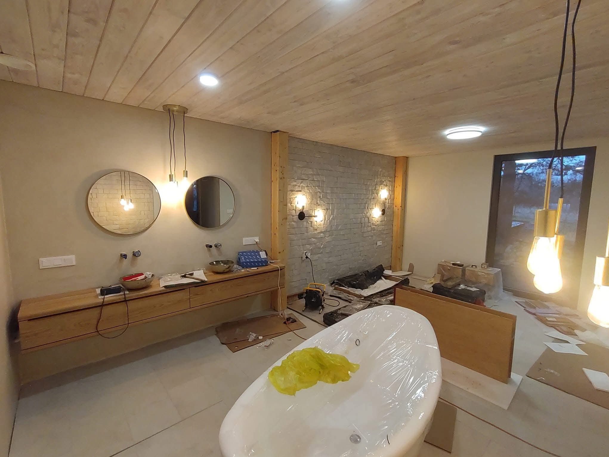 Wnętrze łazienki w trakcie prac wykończeniowych: drewniana zabudowa umywalki z okrągłymi lustrami, wanna zabezpieczona folią, sufit z drewnianych paneli, oświetlenie.