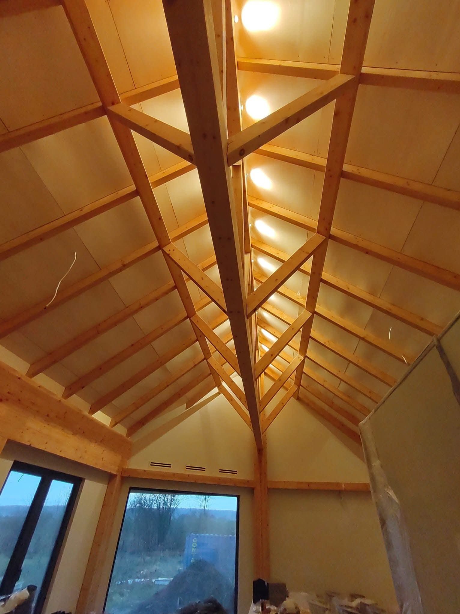 Drewniana konstrukcja dachu z oświetleniem punktowym, widok z dołu. Elementy wykończenia wnętrza w surowym stanie, duże okna z widokiem na zewnątrz.