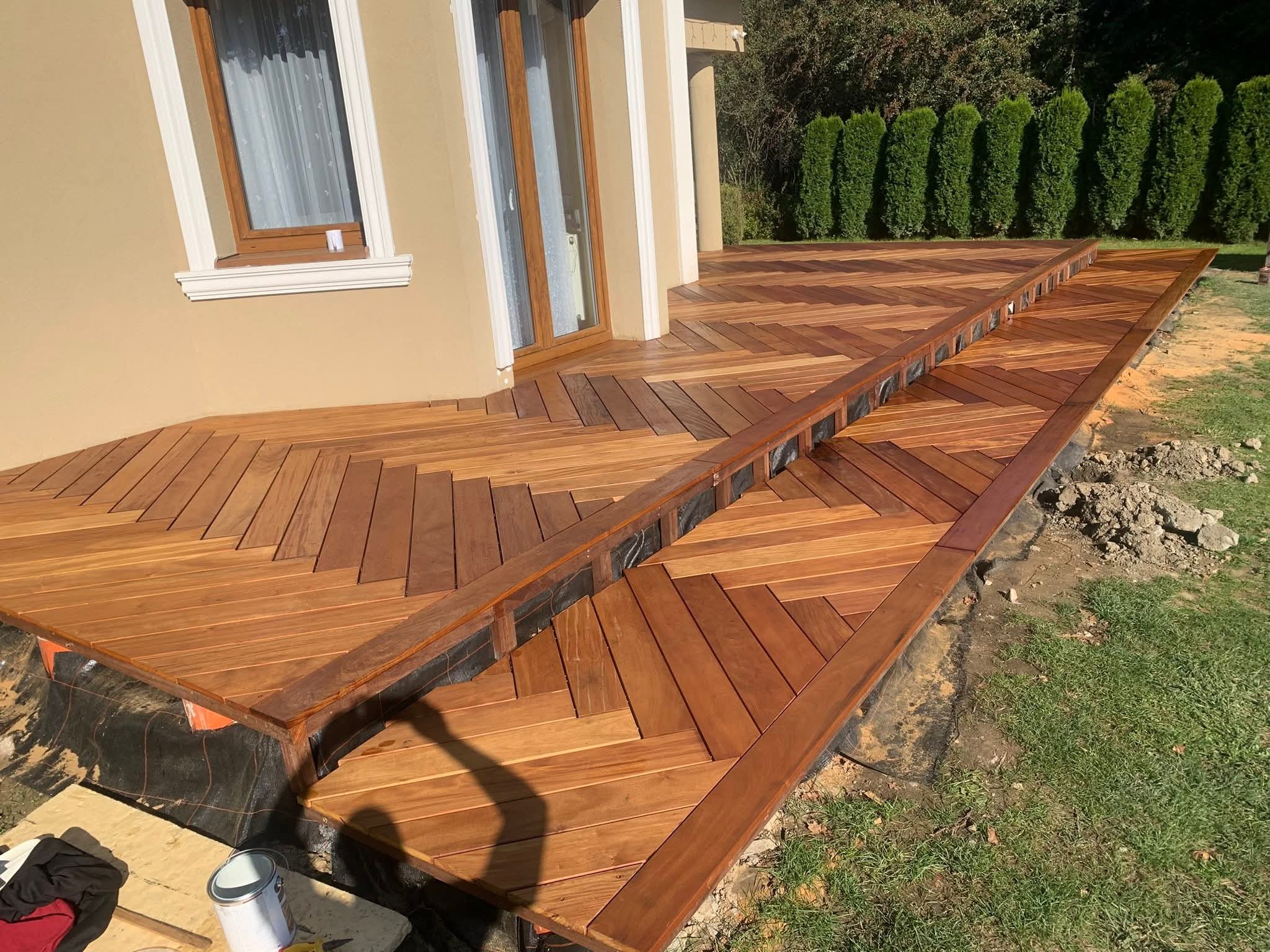 Drewniany taras z desek ułożonych w jodełkę, prowadzący do drzwi domu. Widoczne schody i fragment ogrodu. Deski w ciepłym odcieniu brązu, świeżo po impregnacji.