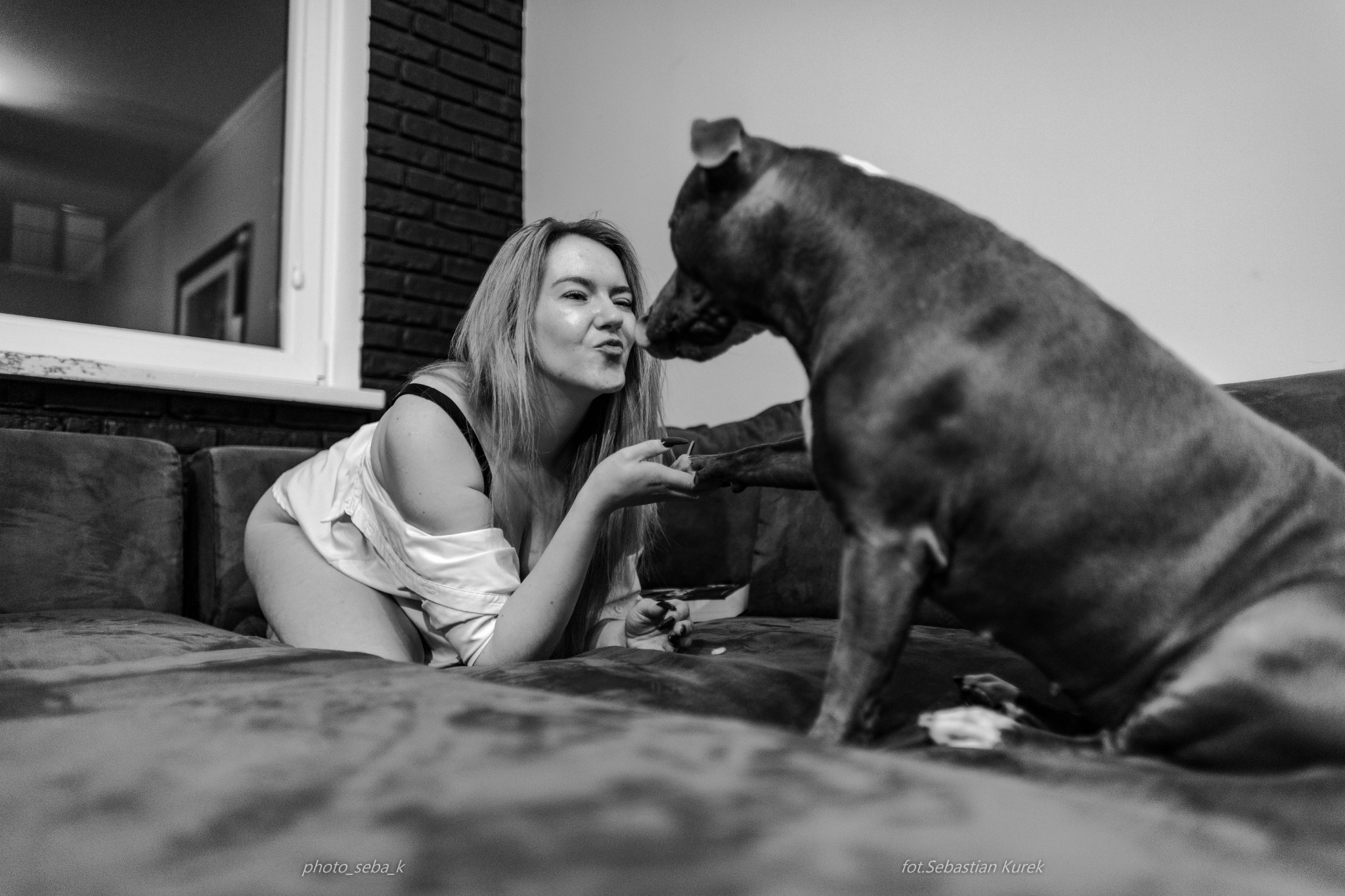 Czarno-białe zdjęcie kobiety w bieliźnie i rozpiętej koszuli, leżącej na kanapie i podającej łapę psu. Ujęcie z bliska, z widocznym oknem w tle.