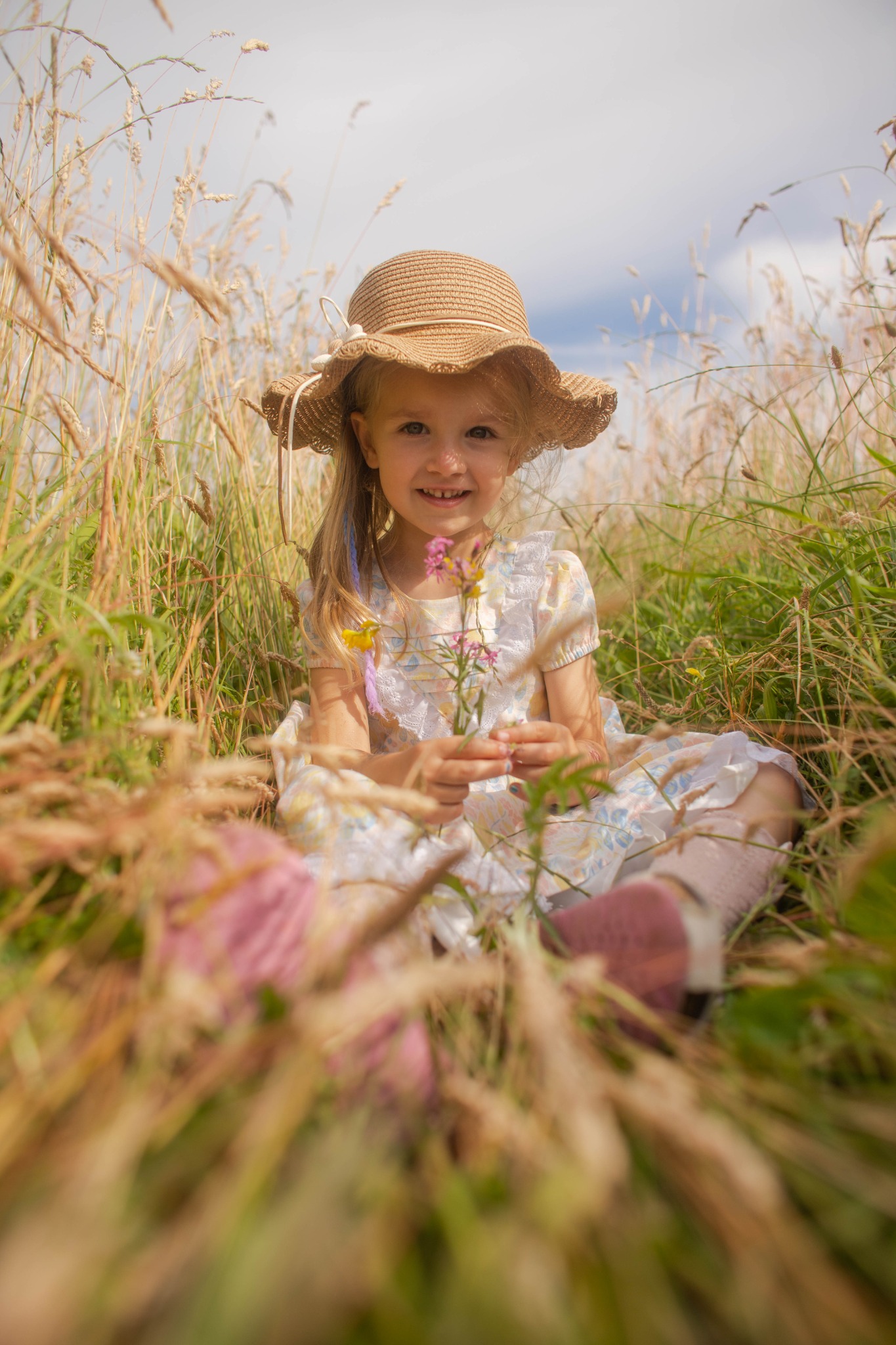 Uśmiechnięta dziewczynka w słomkowym kapeluszu siedzi w trawie, trzymając mały bukiecik polnych kwiatów. Naturalne światło, miękkie kolory, sielski klimat.
