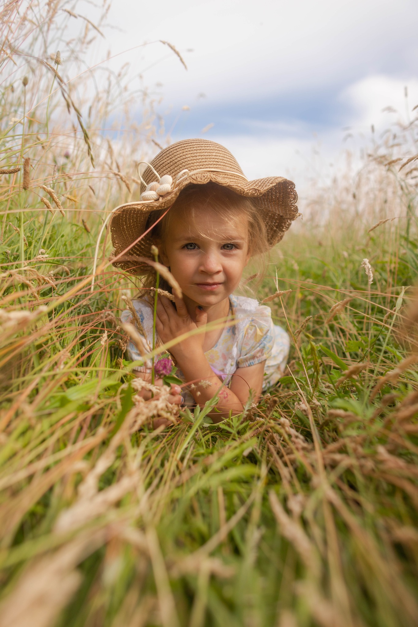 Dziewczynka w słomkowym kapeluszu leży na łące, otoczona wysoką trawą. Uśmiechnięta, patrzy w obiektyw. Naturalne światło, miękkie kolory, plenerowa sesja.