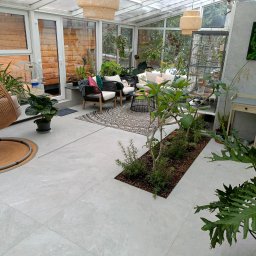 Help Projekt - Jasna oranżeria z szarymi płytkami podłogowymi, rattanowym fotelem wiszącym i roślinami doniczkowymi. Widoczne meble wypoczynkowe i dekoracje.