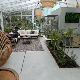 Help Projekt - Jasna oranżeria z szarymi płytkami podłogowymi, rattanowymi lampami, sofą, roślinami doniczkowymi i wiszącym fotelem, tworząca przytulną przestrzeń relaksacyjną.