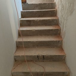 Help Projekt - Surowe, betonowe schody w trakcie remontu, z widocznymi śladami szpachlowania na ścianach i kablem elektrycznym. Ujęcie z dołu, podkreślające pionowość przestrzeni.