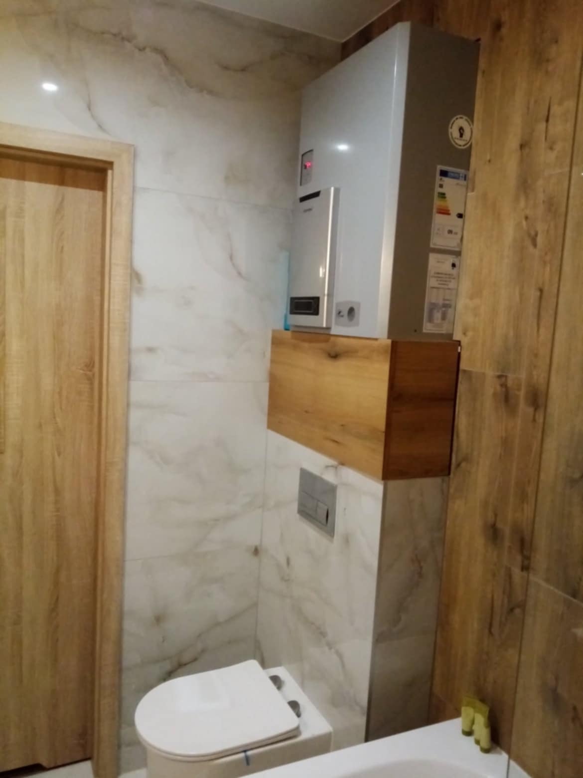 Wykończona łazienka z marmurowymi płytkami, drewnianymi akcentami i zabudowanym geberitem. Wiszący piec gazowy nad szafką. Drzwi z drewnianą okleiną.