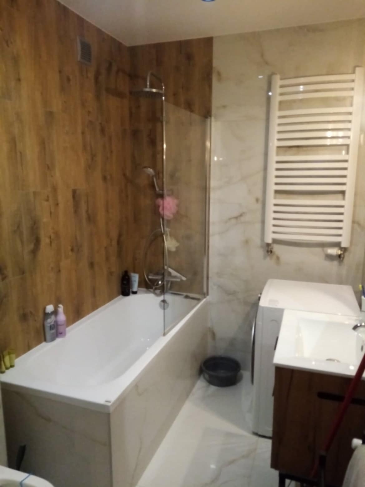 Wykończona łazienka z wanną z parawanem, grzejnikiem drabinkowym i pralką. Ściany wyłożone płytkami imitującymi drewno i marmur. Umywalka z szafką w rogu.