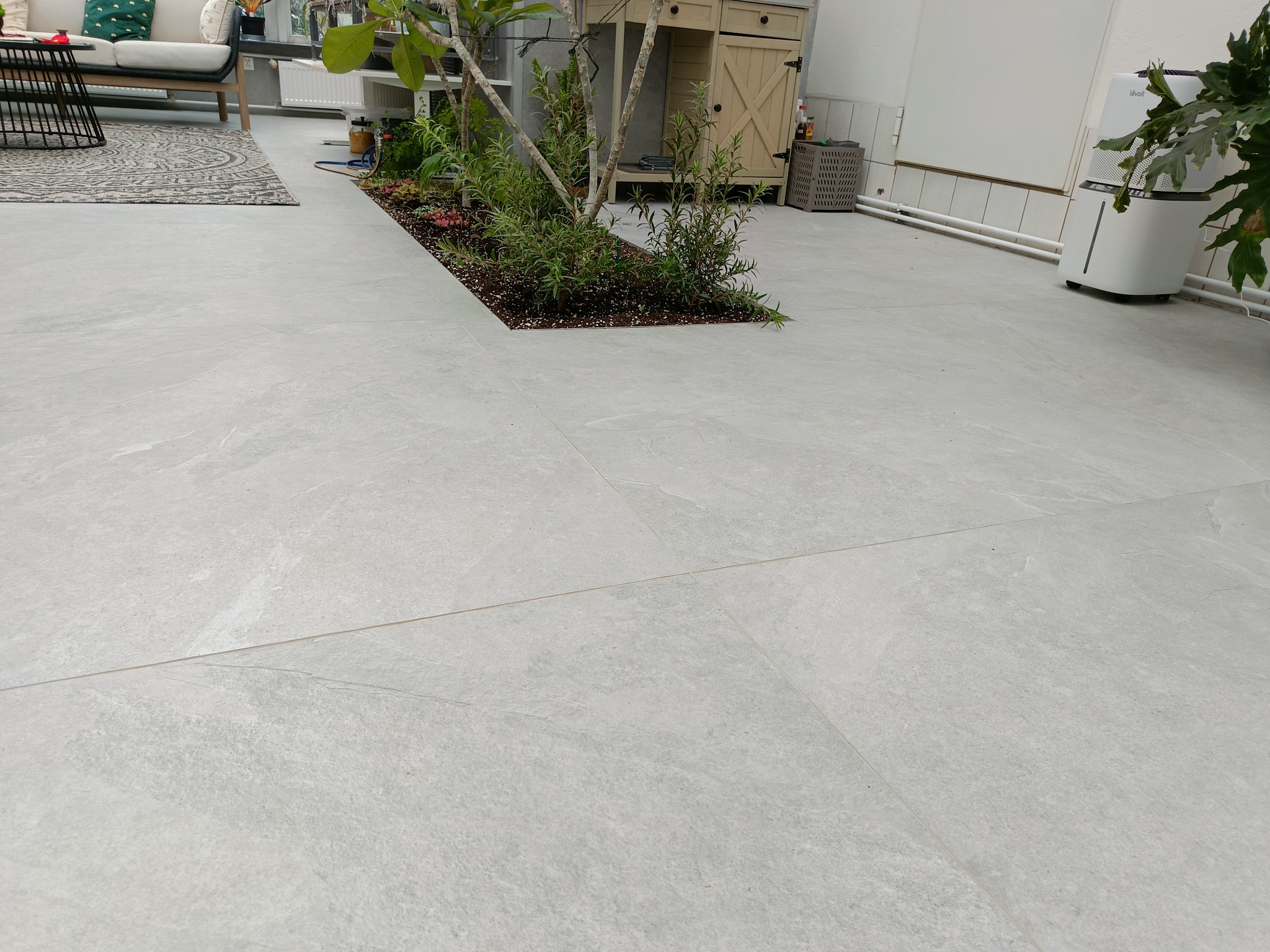 Jasnoszara, matowa podłoga z płytek gresowych w nowoczesnym wnętrzu, z wbudowaną rabatą z roślinami. W tle meble ogrodowe i szafka z drewnianymi drzwiczkami.