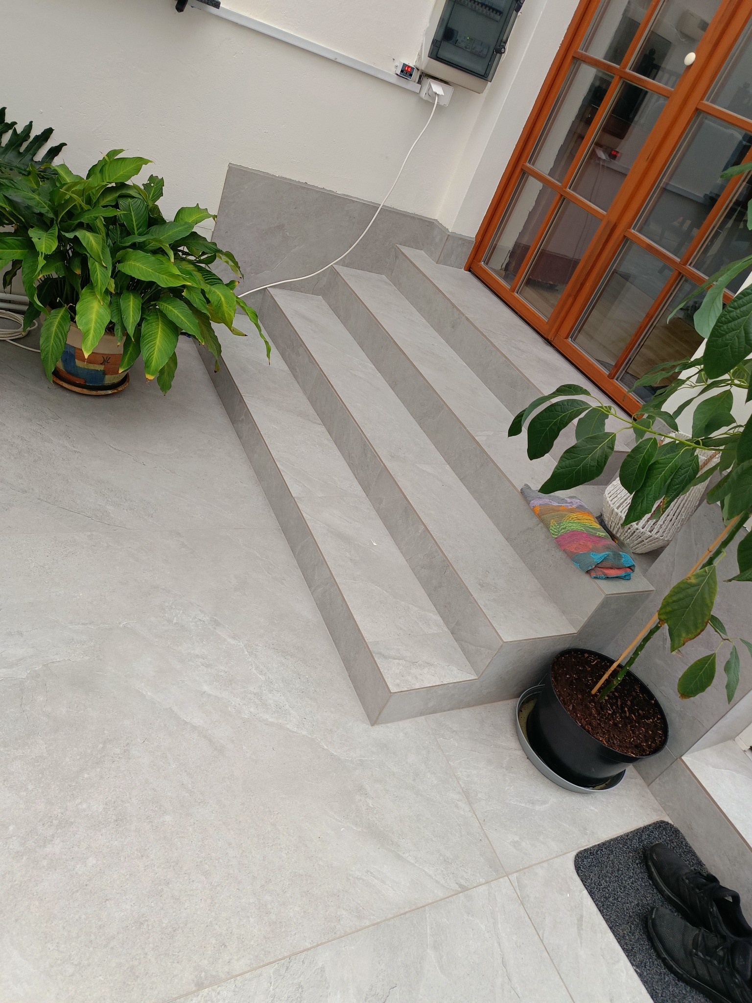 Jasnoszara, wykafelkowana podłoga i schody z roślinami doniczkowymi, prowadzące do drewnianych drzwi z szybami. Widoczna instalacja elektryczna na ścianie.