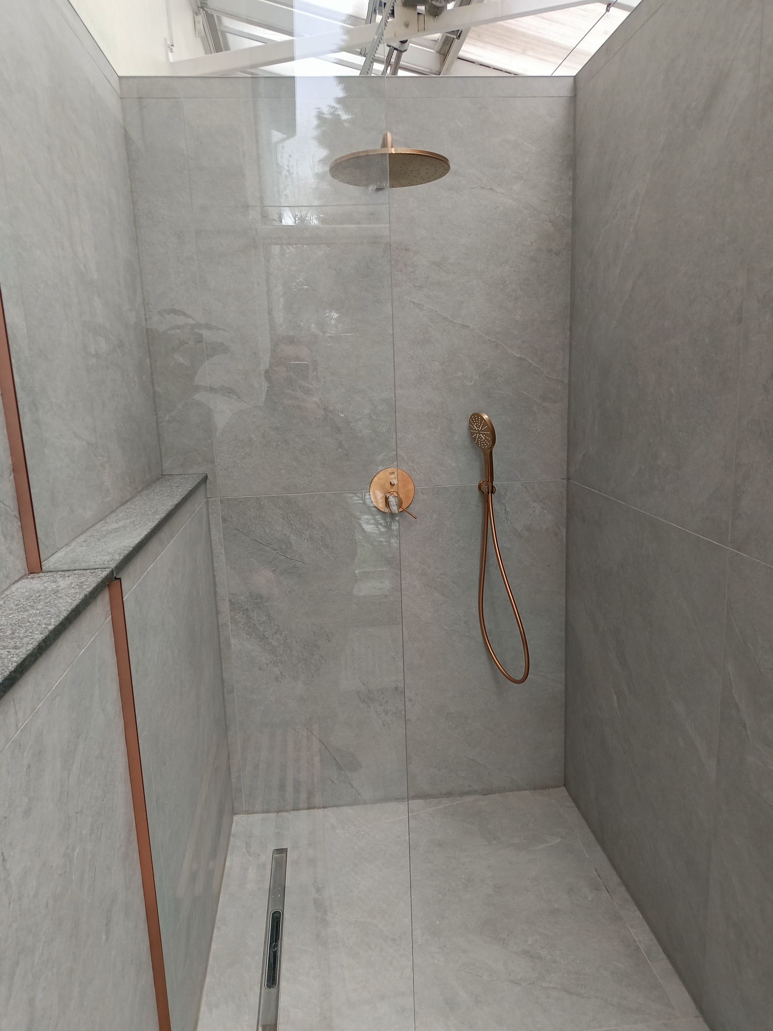 Nowoczesna kabina prysznicowa z szarymi płytkami imitującymi kamień, miedzianą armaturą i odpływem liniowym. Minimalistyczny design z przeszkloną ścianą.