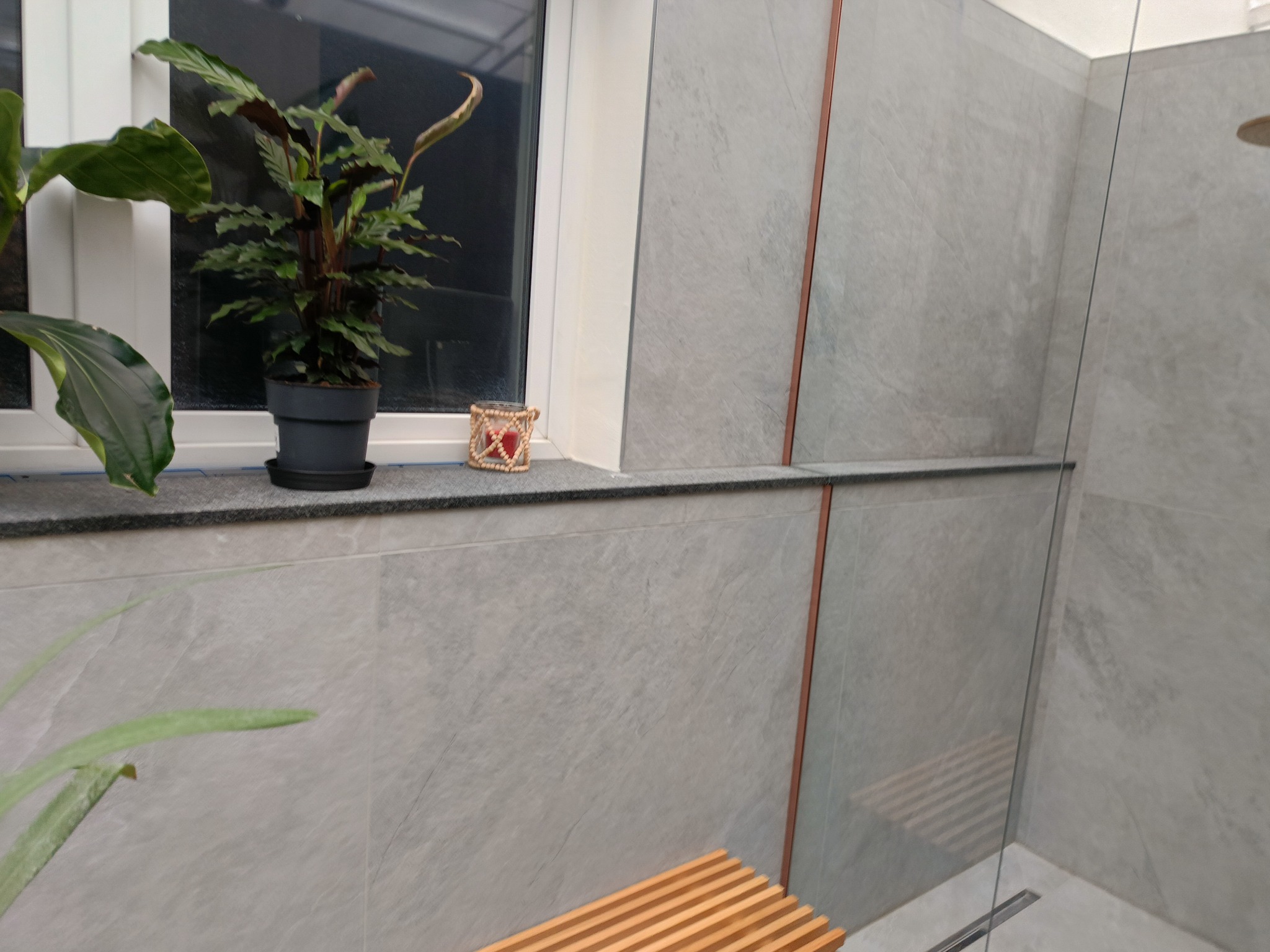 Minimalistyczna łazienka z szarymi płytkami, drewnianym siedziskiem pod prysznicem i roślinami w doniczkach na parapecie okna. Nowoczesny design z akcentami natury.