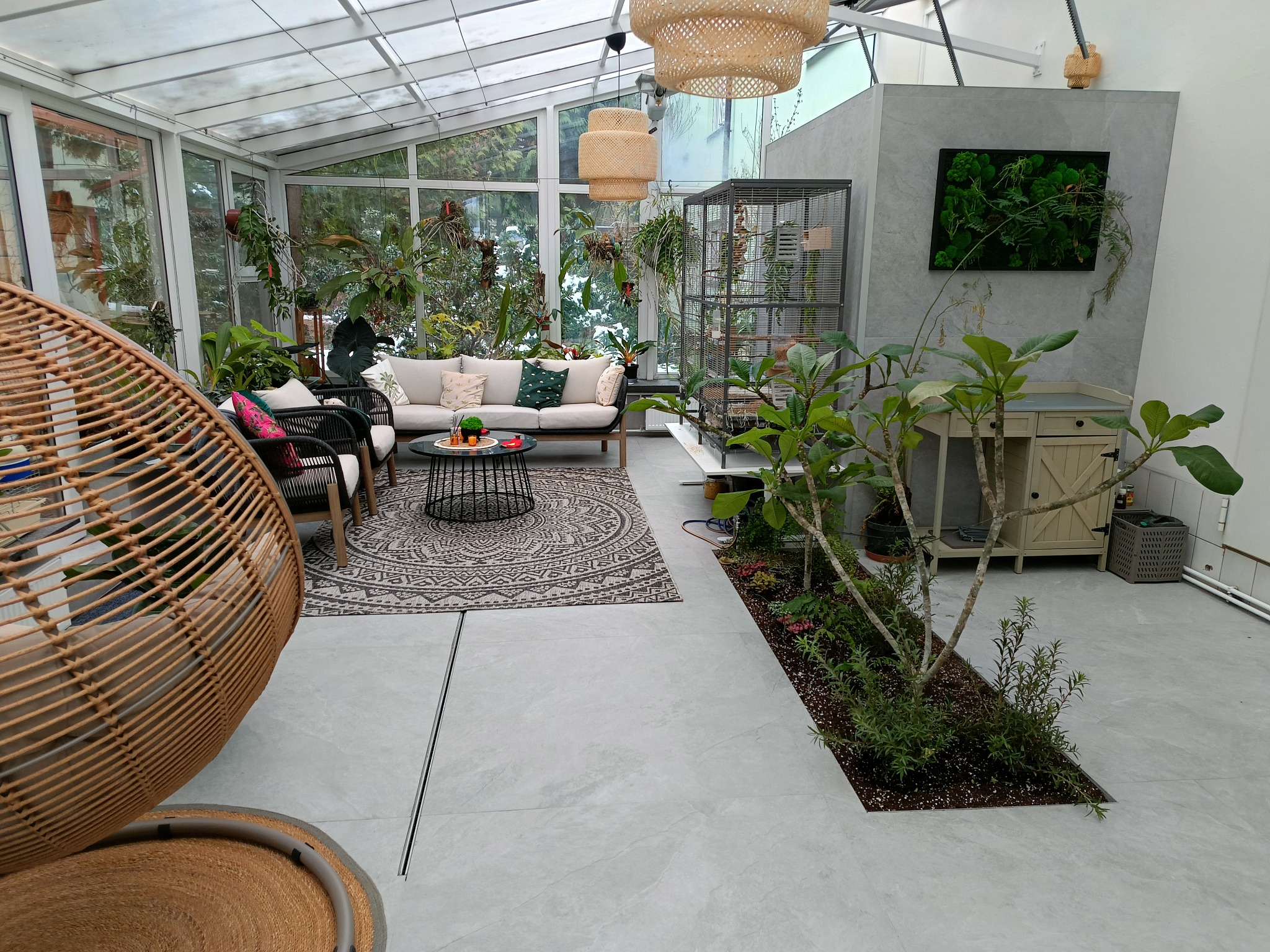 Jasna oranżeria z szarymi płytkami podłogowymi, rattanowymi lampami, sofą, roślinami doniczkowymi i wiszącym fotelem, tworząca przytulną przestrzeń relaksacyjną.