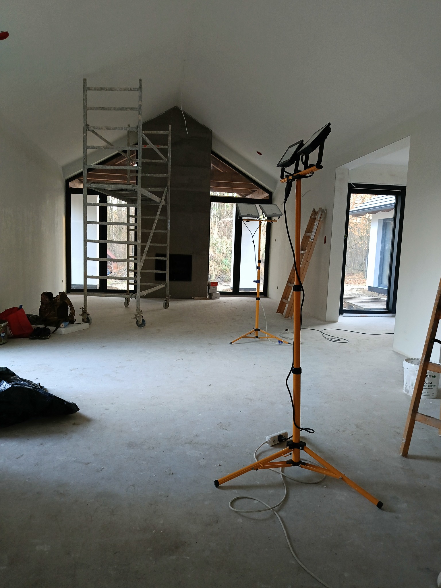 Wnętrze w trakcie wykańczania: surowe ściany, betonowa podłoga, rusztowanie, lampy budowlane. Widoczne okna z czarnymi ramami i komin. Etap prac wykończeniowych.