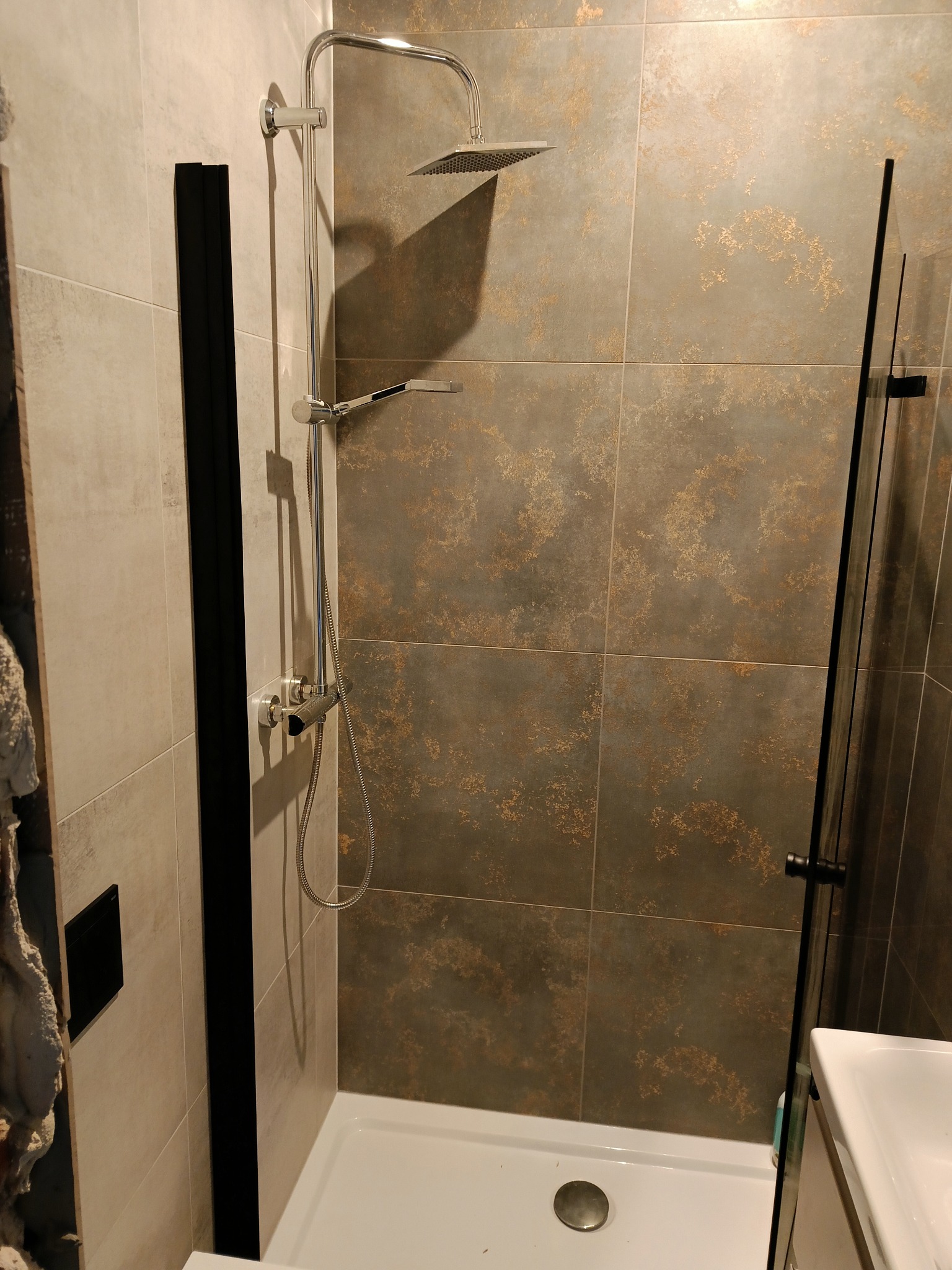 Nowoczesna kabina prysznicowa z deszczownicą i szklaną ścianką, wykończona płytkami w odcieniach szarości i brązu z delikatnym wzorem. Widoczny biały brodzik i fragment umywalki.