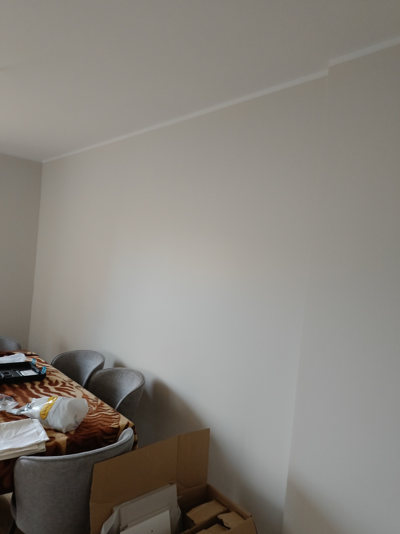 Jasna ściana po malowaniu, widoczna listwa przysufitowa. Częściowo widoczny stół z krzesłami i kartonowe pudła w rogu pokoju.