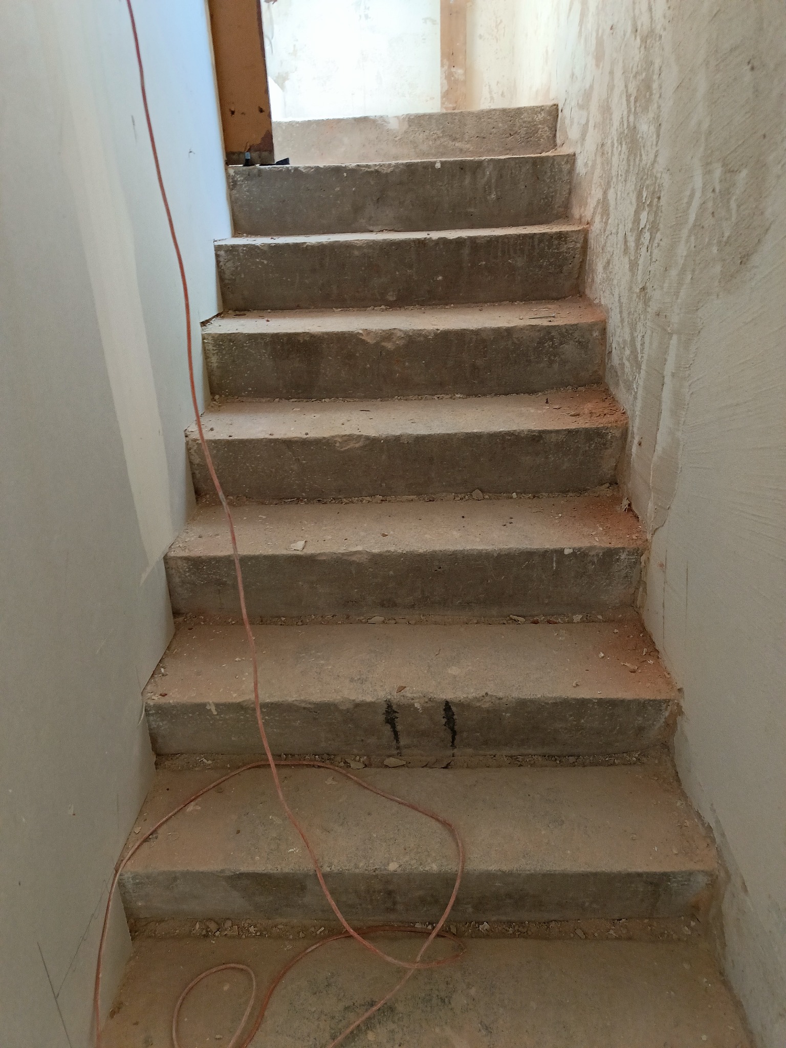 Surowe, betonowe schody w trakcie remontu, z widocznymi śladami szpachlowania na ścianach i kablem elektrycznym. Ujęcie z dołu, podkreślające pionowość przestrzeni.