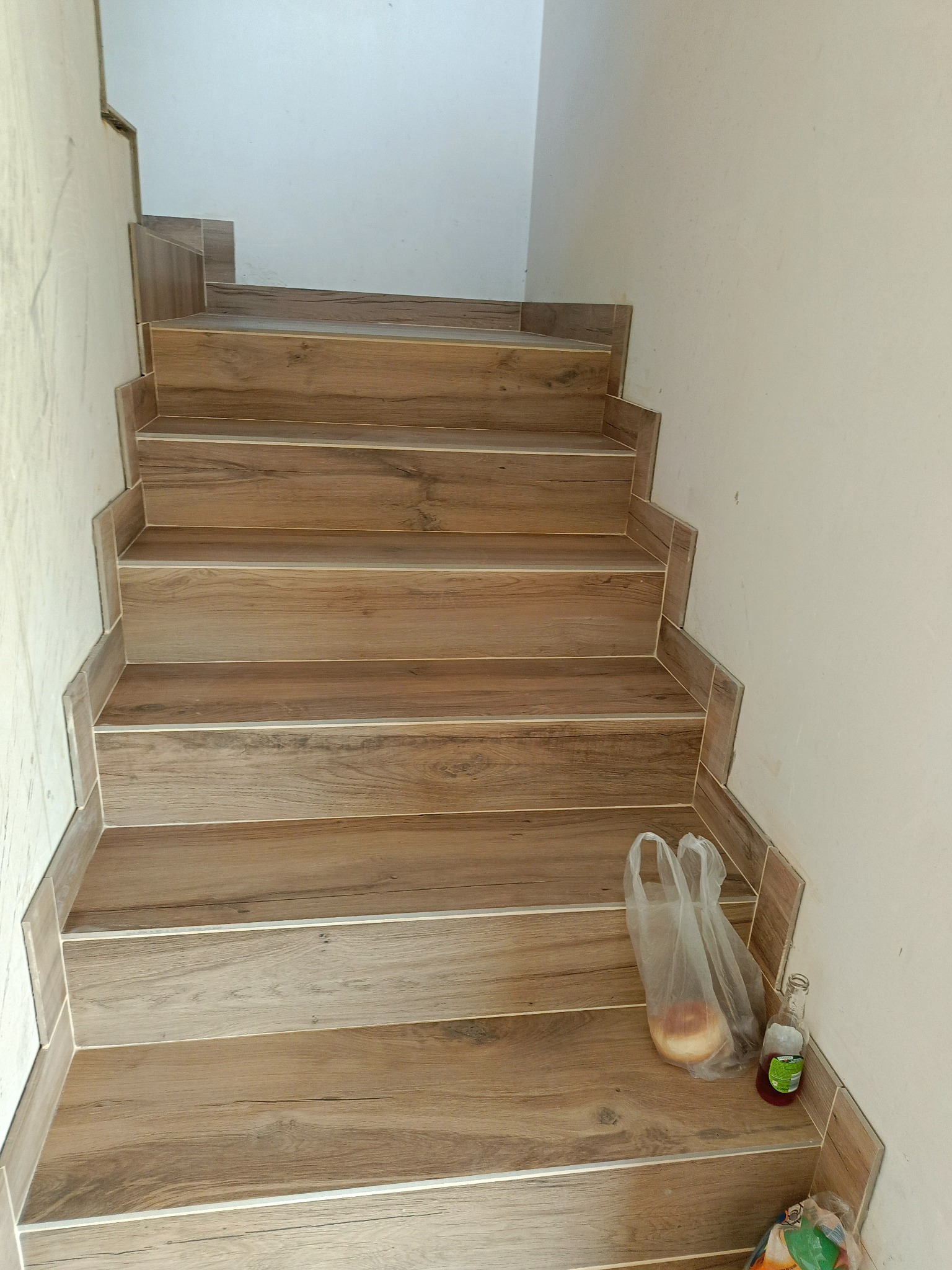Schody wyłożone płytkami imitującymi drewno, z widocznymi detalami fug i krawędzi. Na jednym ze stopni leżą torby z zakupami i butelka, co dodaje realizmu.
