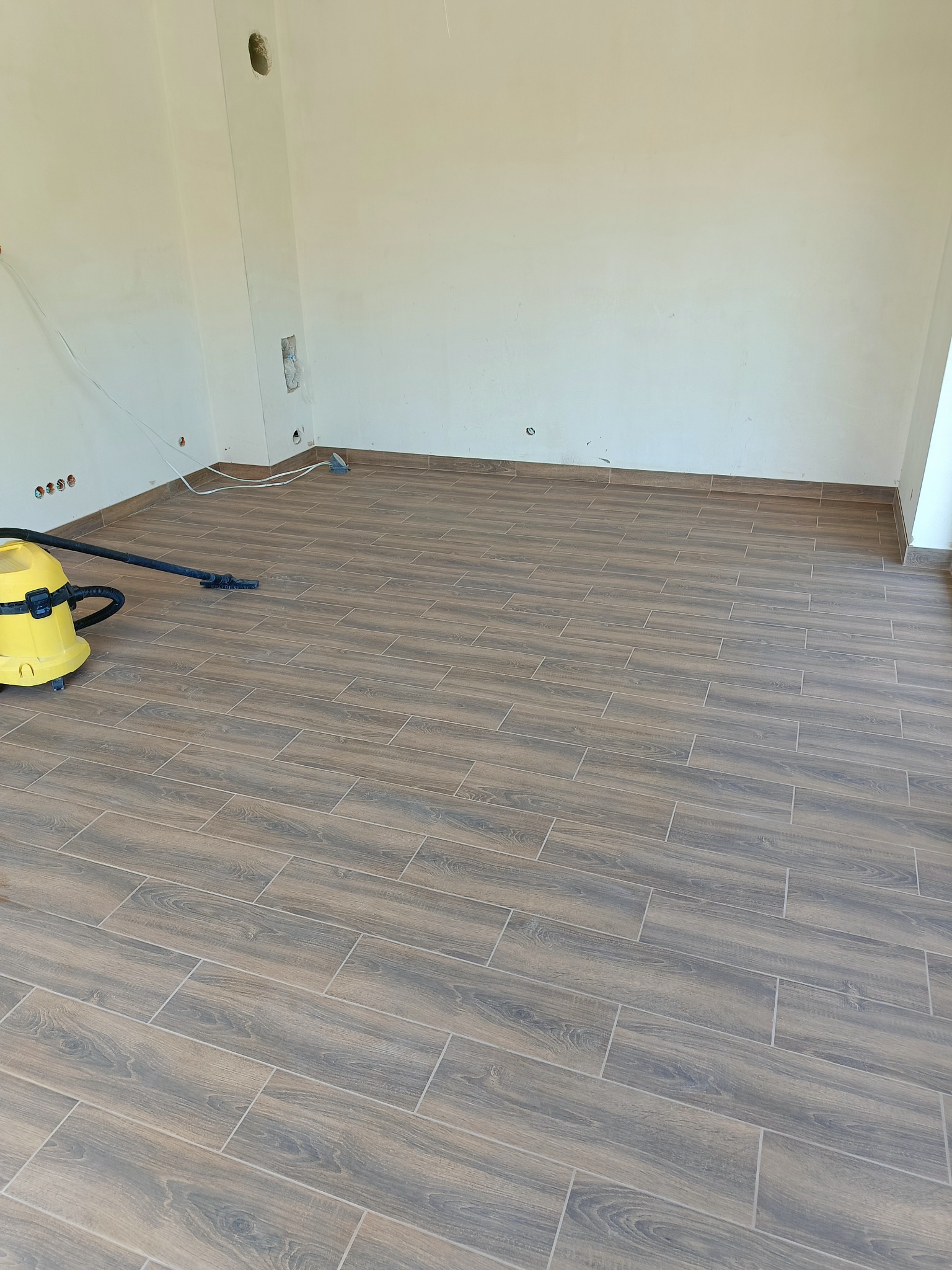 Ułożona podłoga z płytek imitujących drewno w jasnym pomieszczeniu. Żółty odkurzacz przemysłowy w rogu. Widoczne gniazdka elektryczne i otwory w ścianie.