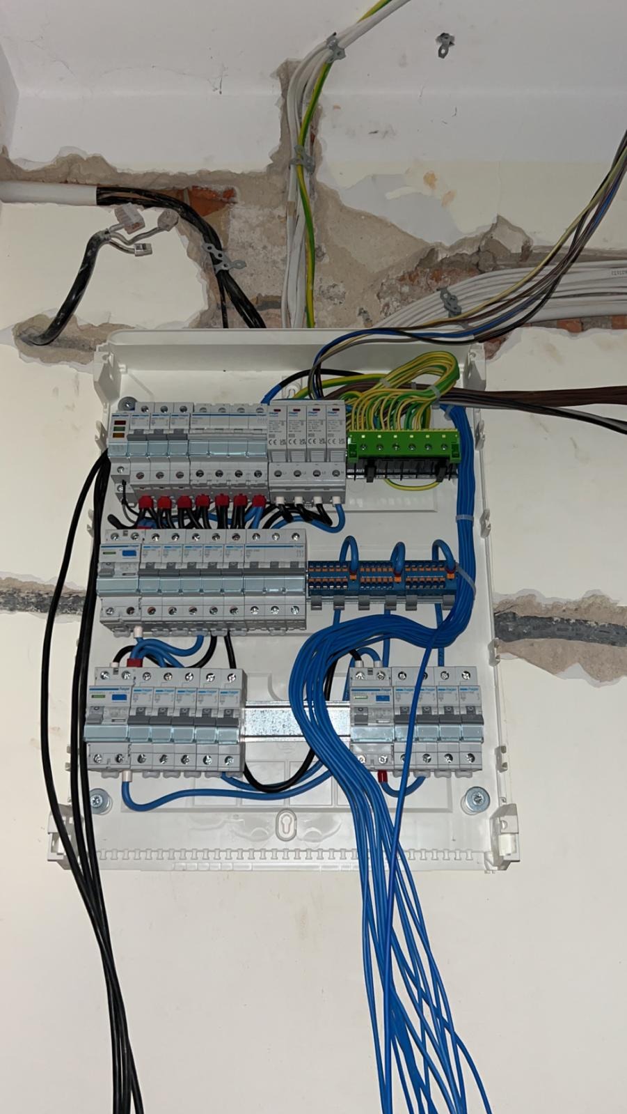 Rozdzielnica elektryczna z podłączonymi przewodami w kolorach: niebieskim, czarnym, żółto-zielonym. Widoczne zabezpieczenia i listwy zaciskowe. Ściana w tle z widocznymi śladami po kuciu.