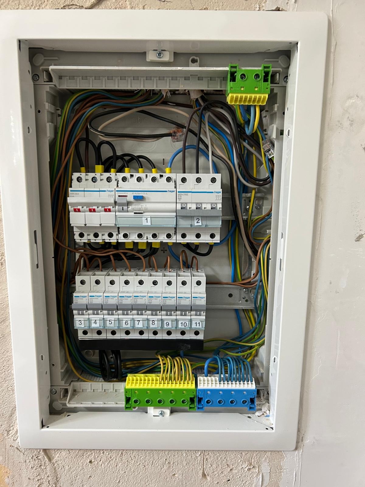 Rozdzielnica elektryczna Hager z zainstalowanymi bezpiecznikami i przewodami w kolorach: brązowym, niebieskim, żółto-zielonym. Widoczne listwy zaciskowe.