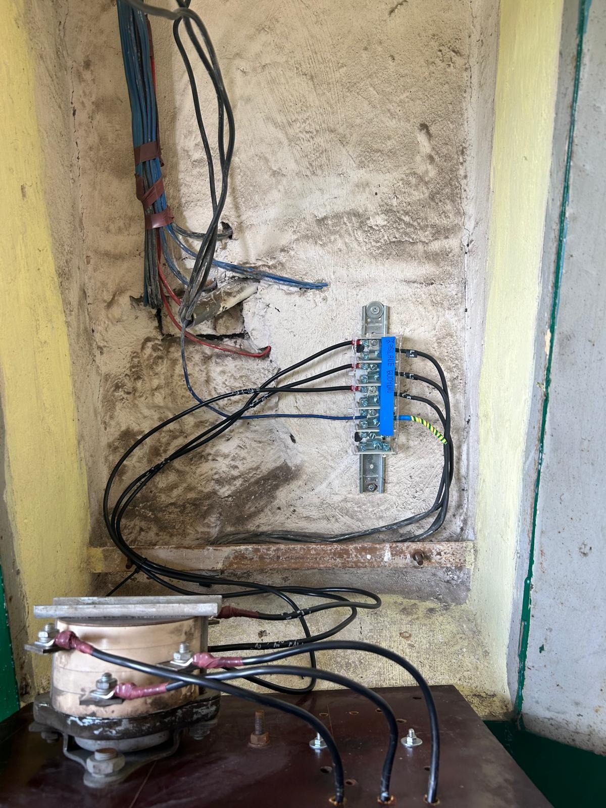 Rozdzielnica elektryczna z przewodami w starej, pomalowanej na żółto wnęce. Widoczne złącza, kable w izolacji i elementy transformatora. Prace instalacyjne w budynku.