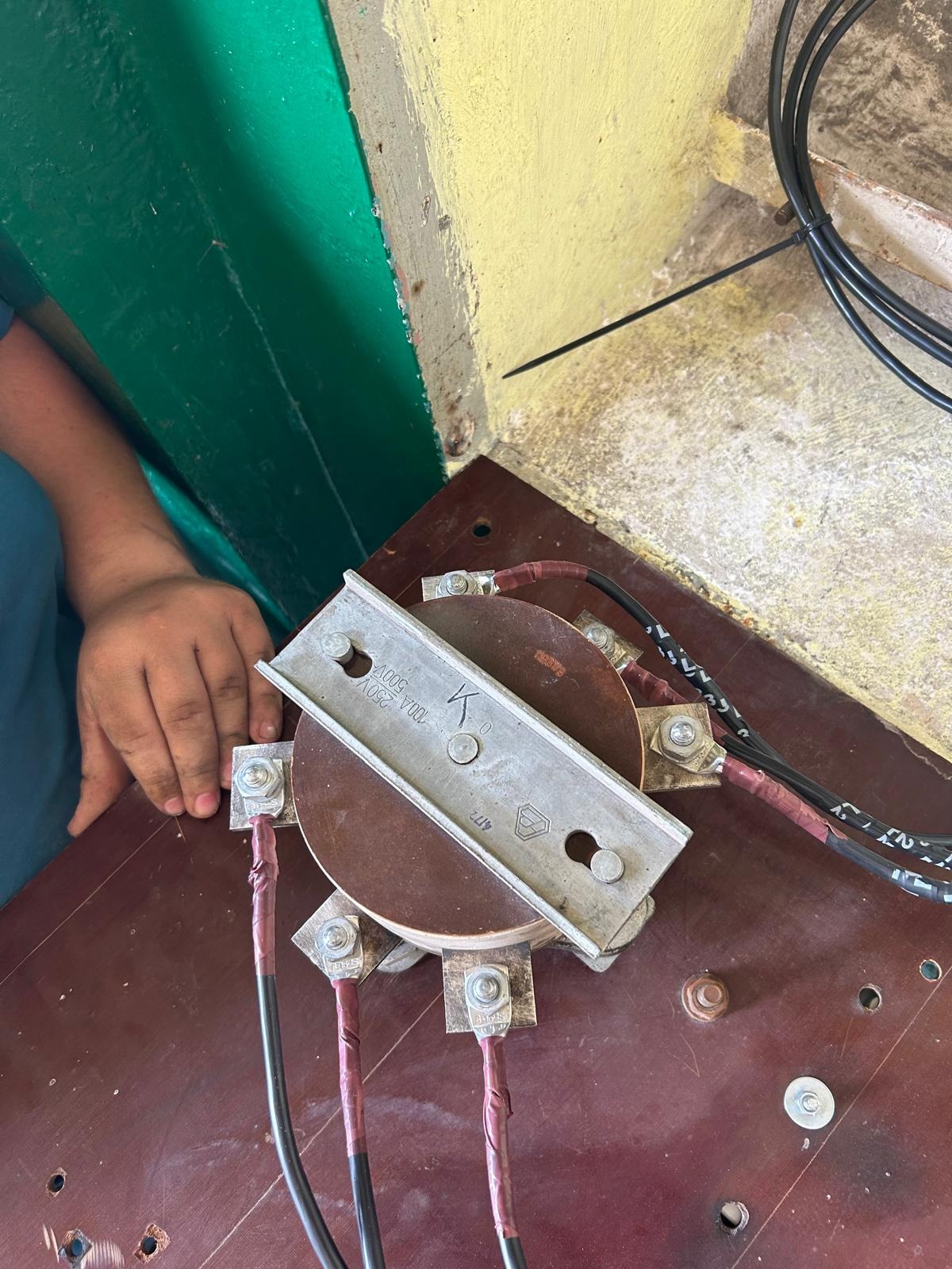 Podłączenie przewodów do transformatora z użyciem metalowej listwy. Widoczne śruby, nakrętki i izolacja przewodów. Praca w toku, widoczna dłoń.