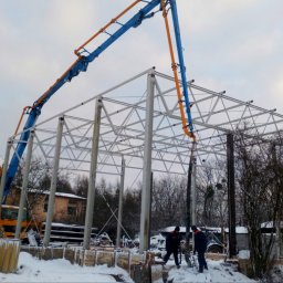 M-BET Częstochowa - Zimowy dzień na budowie: pompa do betonu z niebiesko-pomarańczowym ramieniem wylewa mieszankę do szalunków pod konstrukcję stalową hali. Widoczni pracownicy.