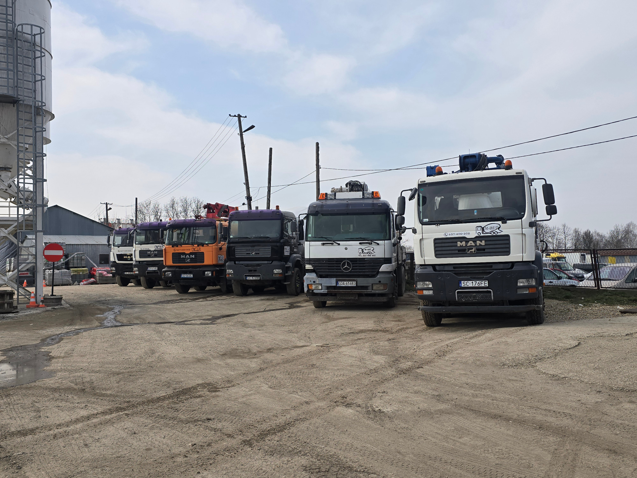 Flota pojazdów ciężarowych firmy z Lubojenki, w tym MAN i Mercedes, zaparkowana na placu. Widoczny silos betoniarni w tle. Różne kolory kabin.
