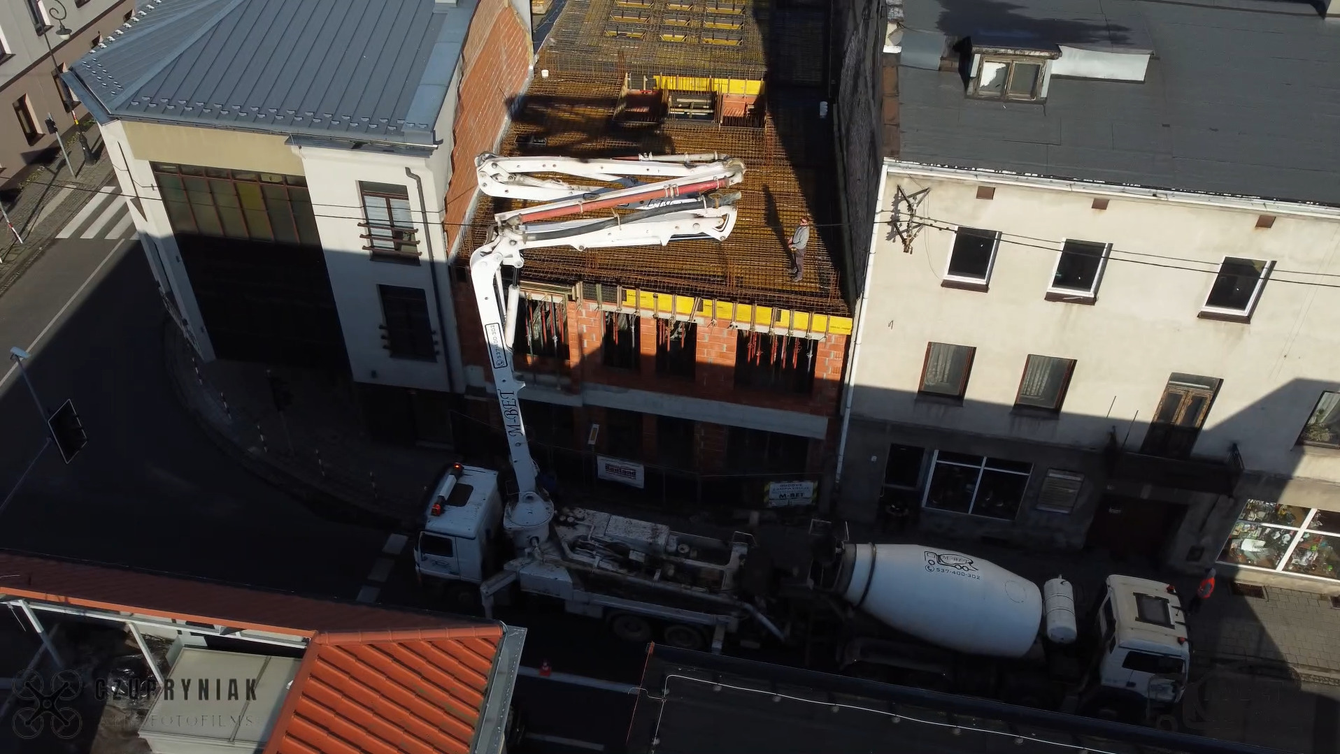 Z góry: pompa do betonu podaje mieszankę na budowę. Widoczny mikser na podwoziu ciężarówki i ramię pompy nad budynkiem w trakcie wylewania stropu.