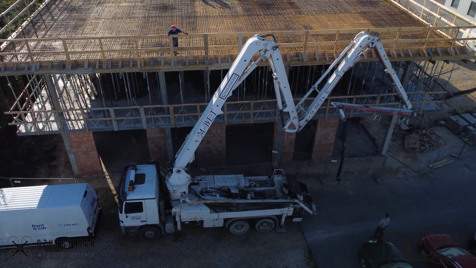 Z góry: pompa do betonu M-BET w akcji na budowie. Widoczny pracownik w kasku na rusztowaniu i częściowo zaparkowane pojazdy. Ujęcie z drona.