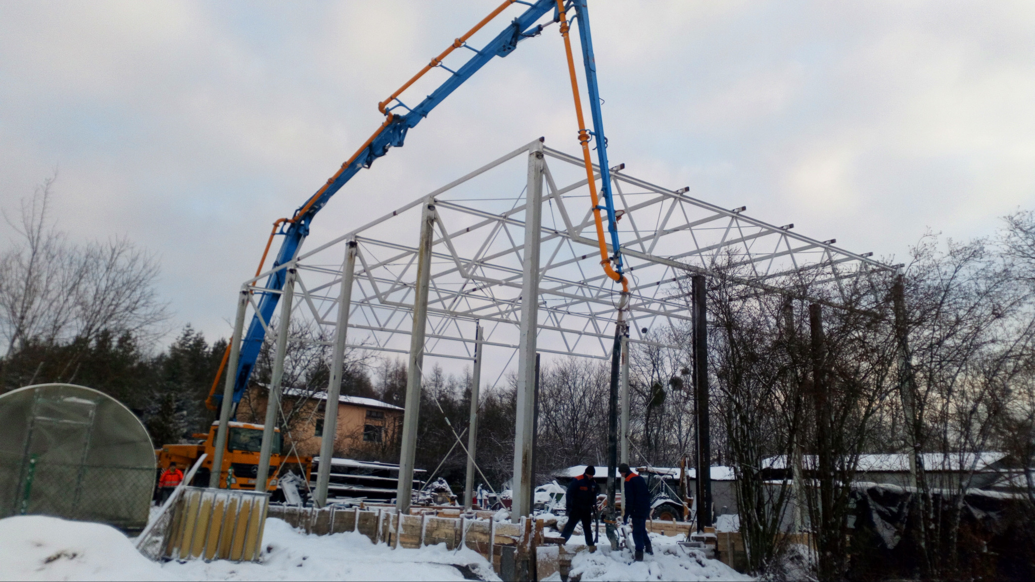 Zimowy dzień na budowie: pompa do betonu z niebiesko-pomarańczowym ramieniem wylewa mieszankę do szalunków pod konstrukcję stalową hali. Widoczni pracownicy.