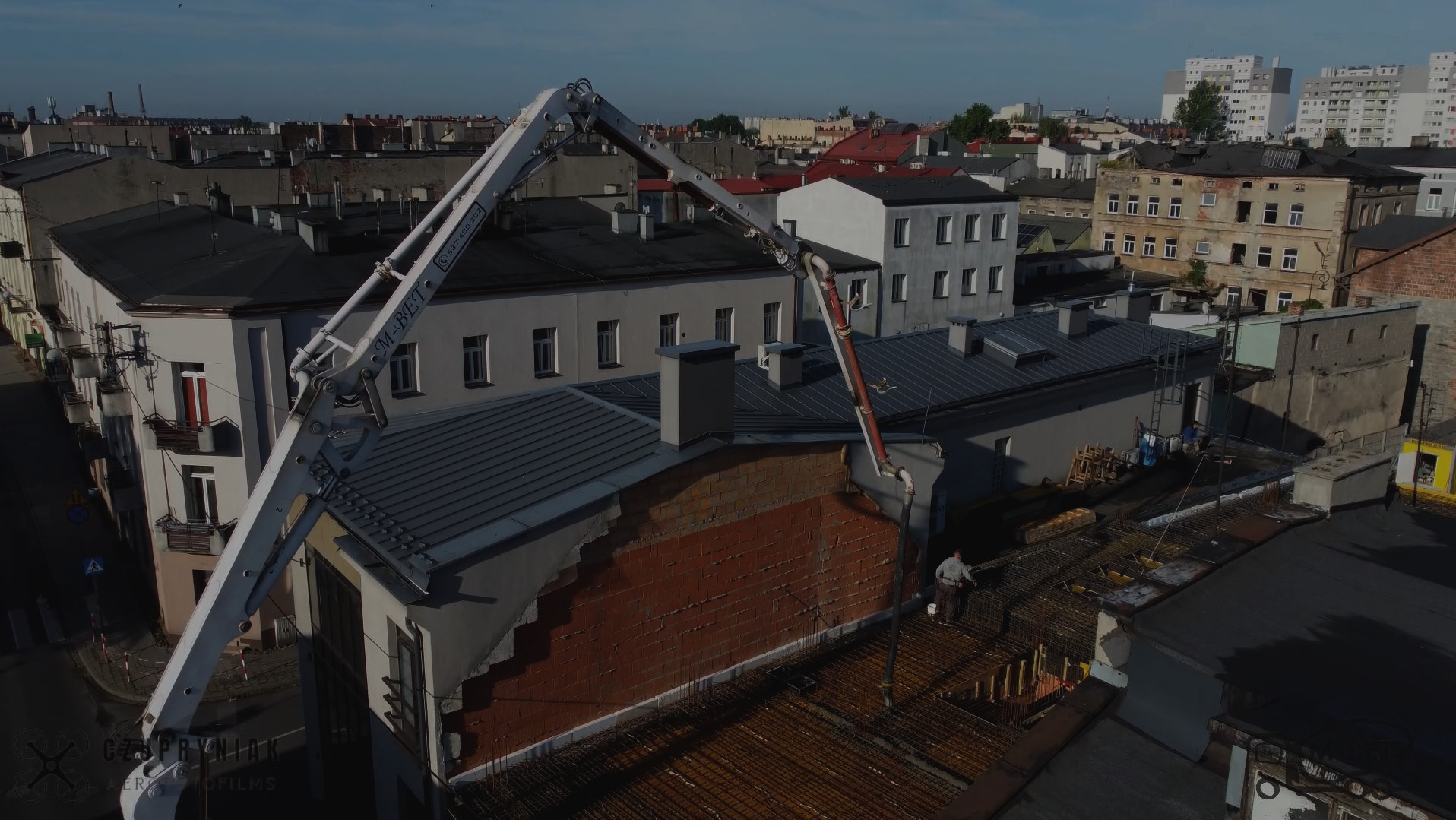 Betonowanie stropu budynku z użyciem pompy do betonu. Widok z góry na zbrojenie i pracownika. W tle zabudowa miejska w słoneczny dzień.