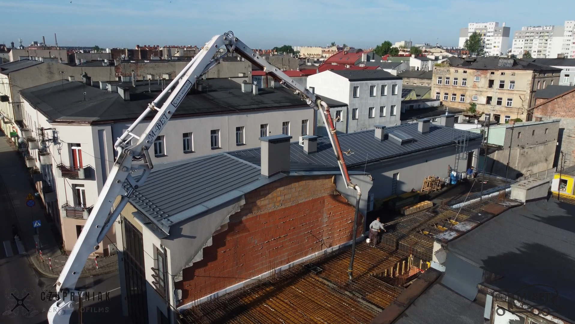 Betonowanie stropu budynku z użyciem pompy do betonu na tle zabudowy miejskiej. Widoczne zbrojenie i pracownik nadzorujący wylewanie betonu.