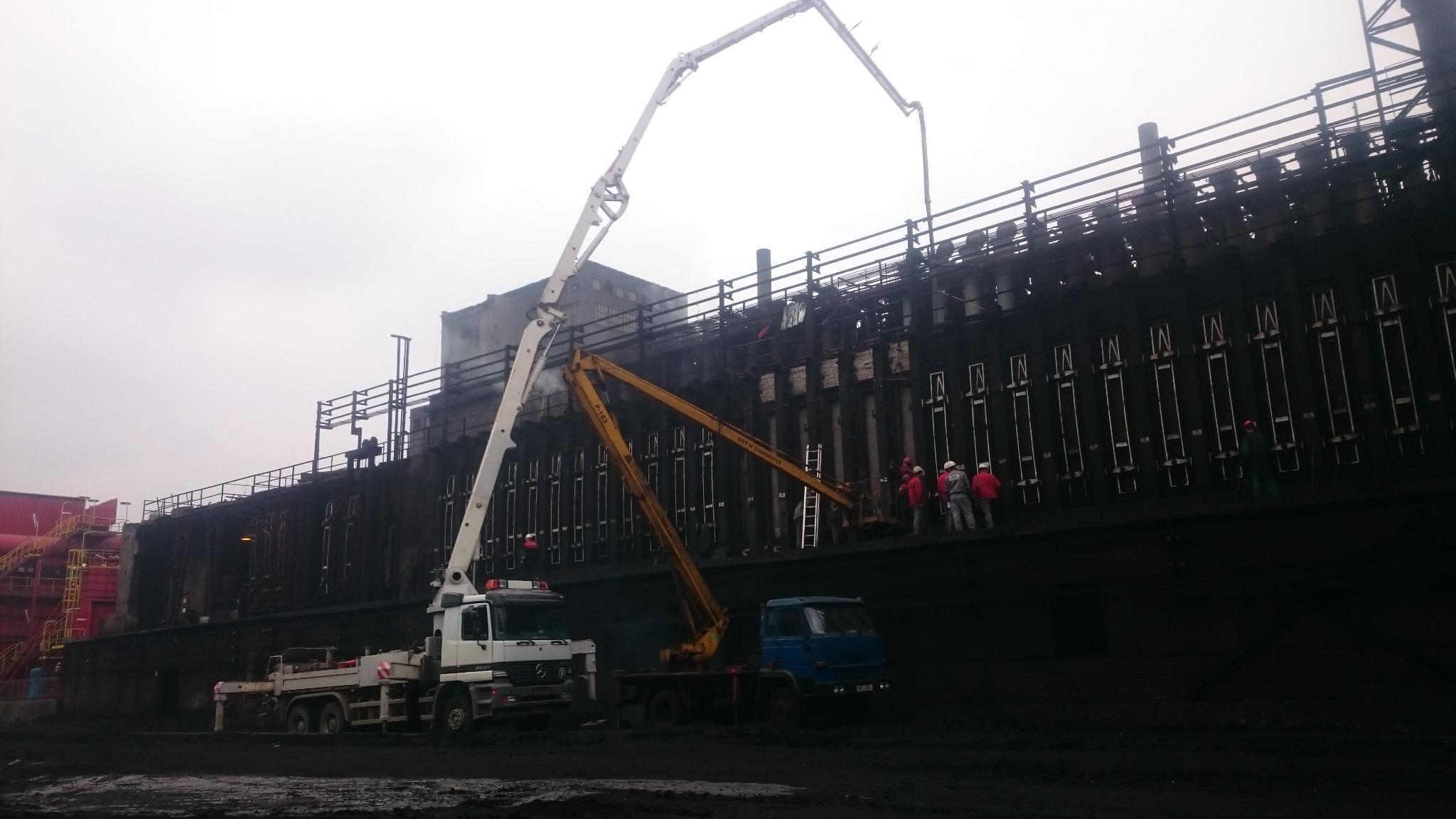 Dwa auto-mikser z pompami do betonu podczas wylewania mieszanki na konstrukcję. Pracownicy w kaskach nadzorują proces. Pochmurne niebo w tle.