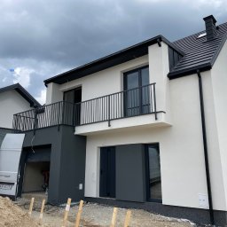 Zbigniew Tyrpula - Nowoczesny dom z balkonem i garażem w trakcie wykończenia elewacji. Widoczny fragment dachu i pracownik w garażu. Biało-szara kolorystyka, pochmurne niebo.