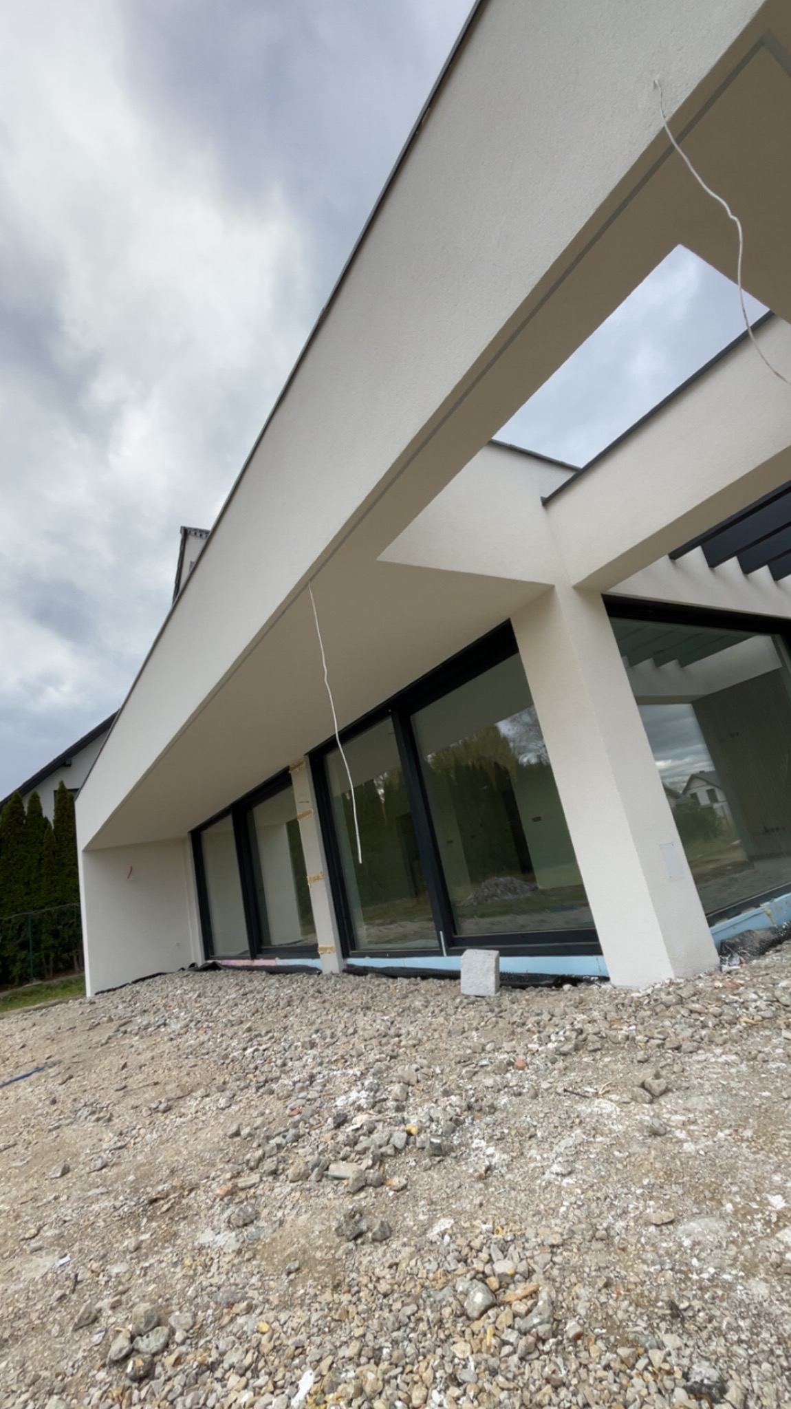 Nowoczesny dom z dużymi oknami i białą elewacją w trakcie wykończenia. Widoczne elementy konstrukcyjne i przygotowanie terenu wokół budynku.
