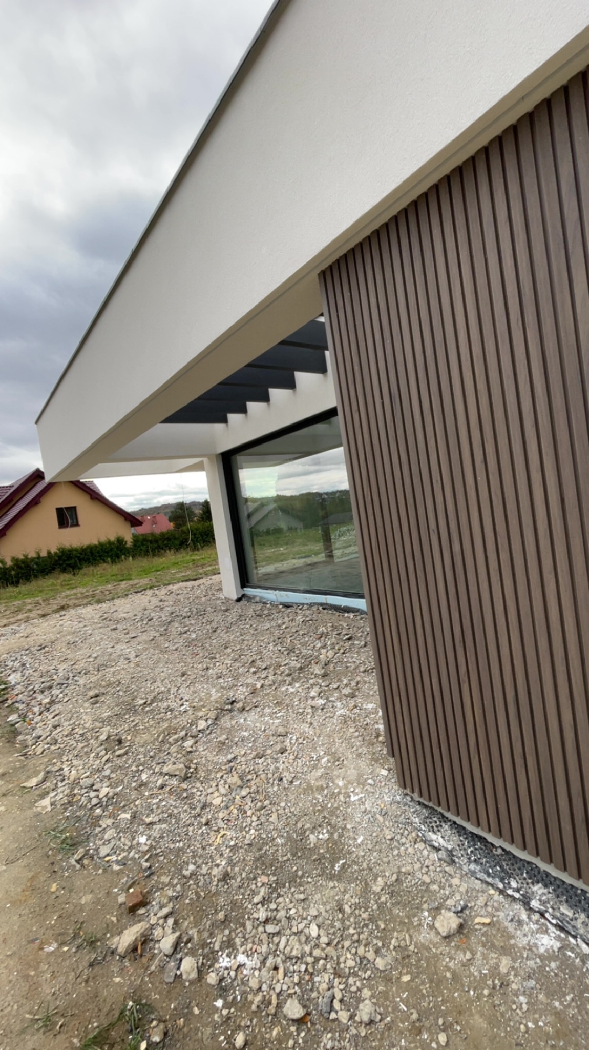 Nowoczesny dom z elewacją z paneli drewnopodobnych i dużym oknem, widok z zewnątrz na żwirowe podłoże i fragment sąsiedniego budynku w tle.