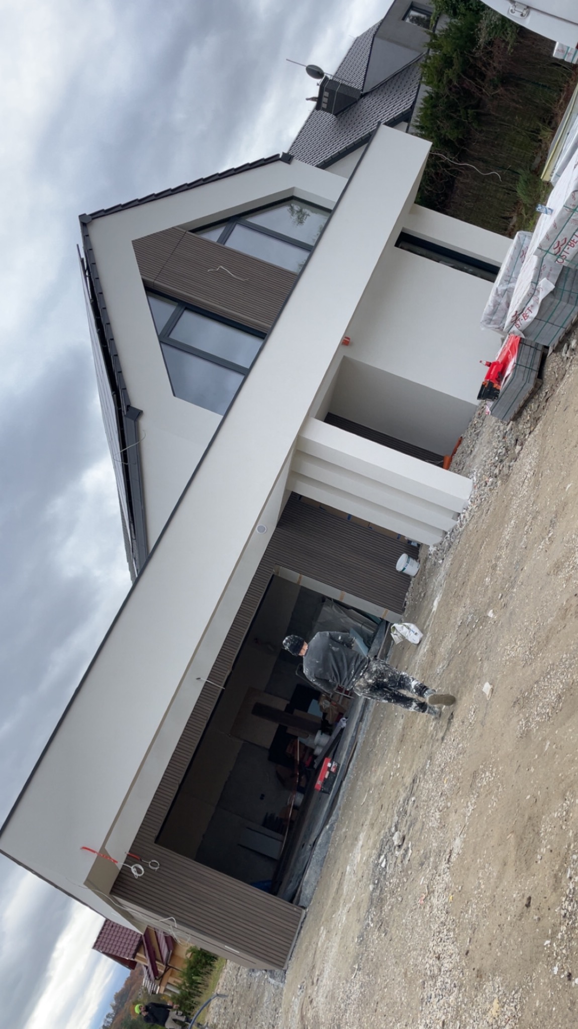 Nowoczesny dom jednorodzinny z białą elewacją i drewnianymi panelami, widok z dołu, pracownik w ubraniu roboczym na pierwszym planie, materiały budowlane obok budynku.