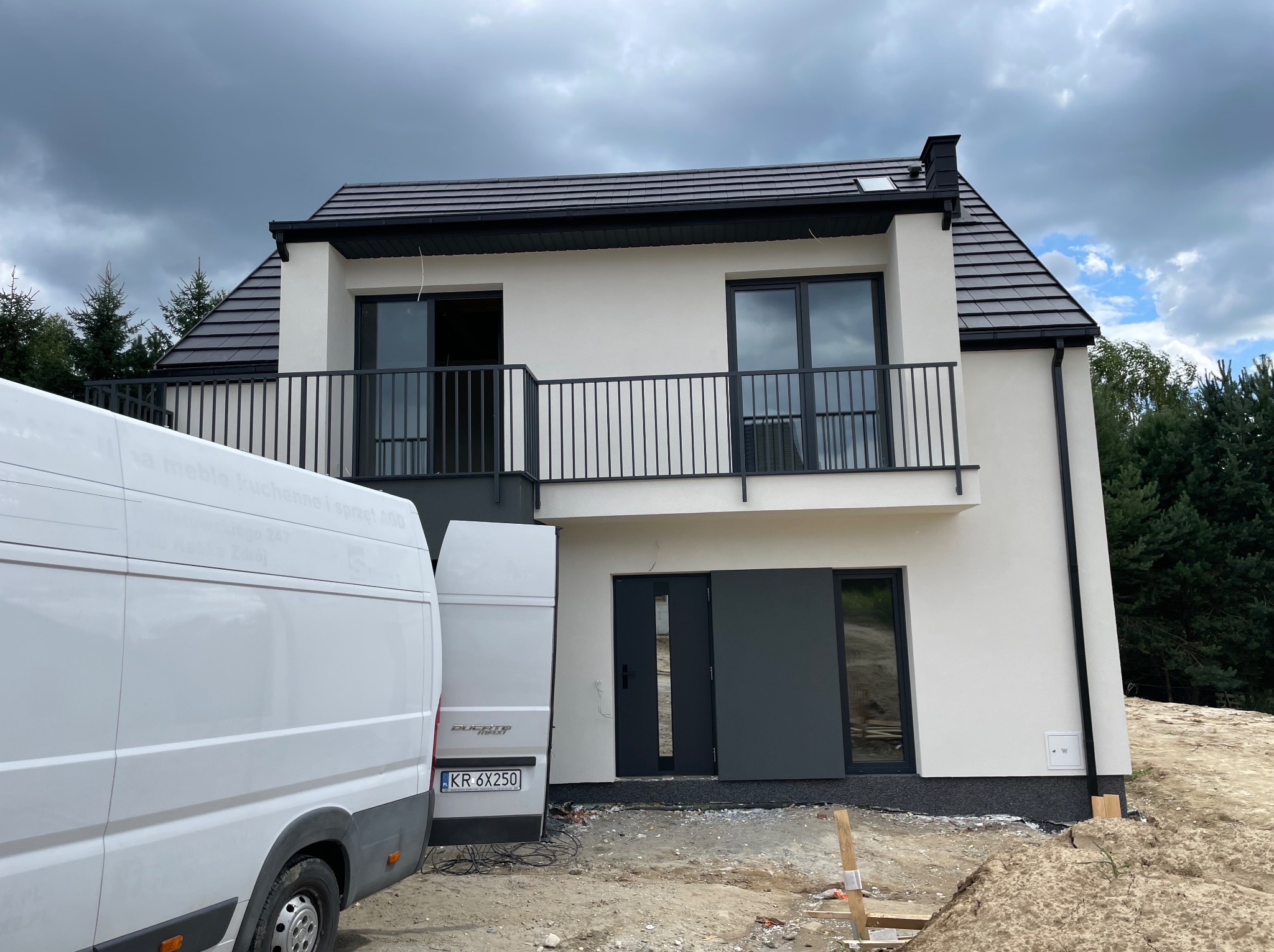 Nowoczesny, dwukondygnacyjny dom z balkonami i ciemną stolarką okienną, świeżo po tynkowaniu, z dwoma samochodami dostawczymi na pierwszym planie.