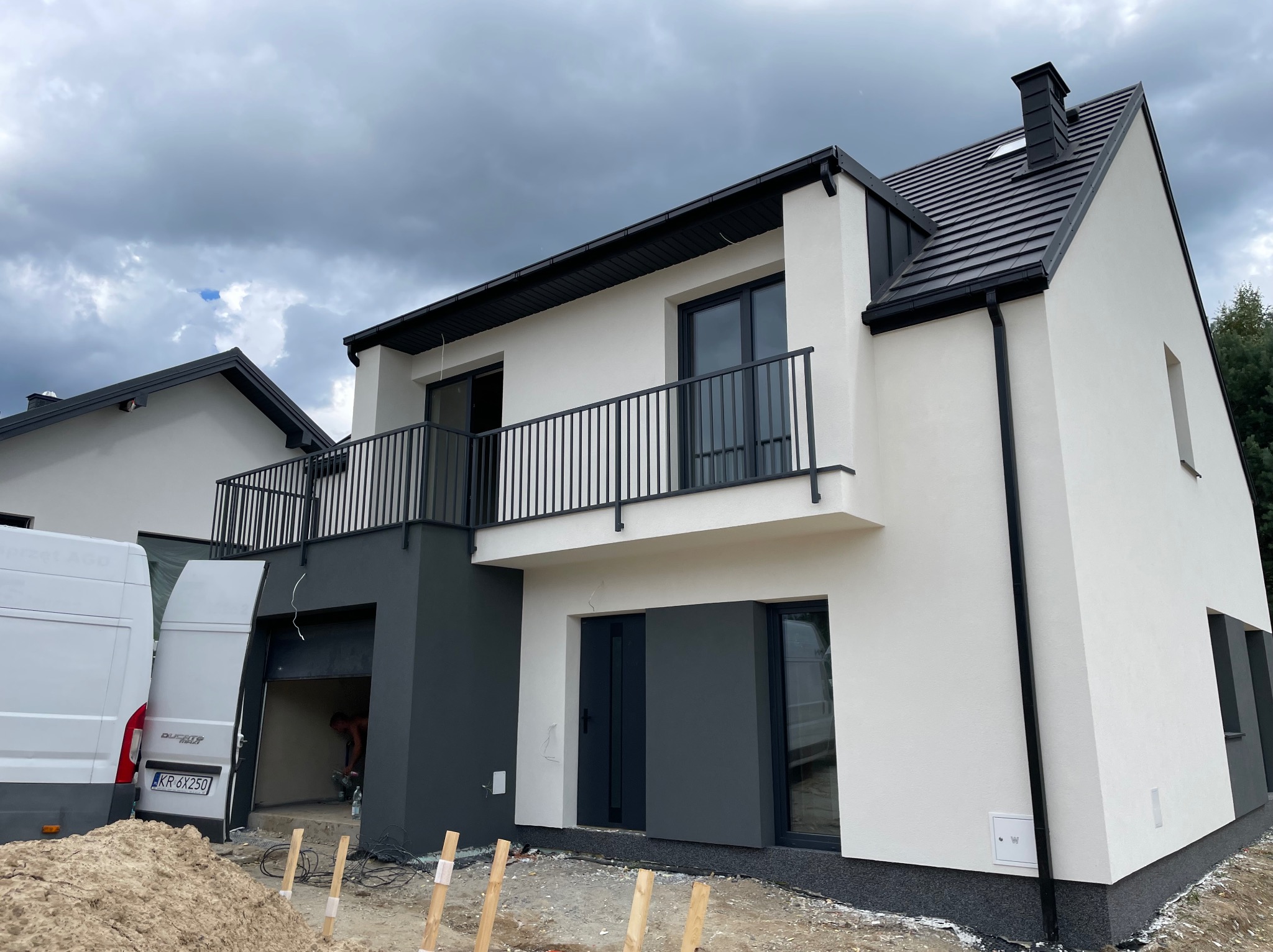 Nowoczesny dom z balkonem i garażem w trakcie wykończenia elewacji. Widoczny fragment dachu i pracownik w garażu. Biało-szara kolorystyka, pochmurne niebo.