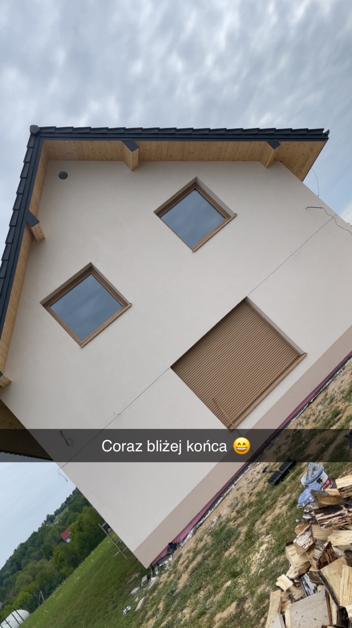 Elewacja domu z oknami w kształcie rombów i roletą zewnętrzną, widok z dołu, ukośny kadr. Widoczne drewniane elementy dachu i stos drewna obok budynku.