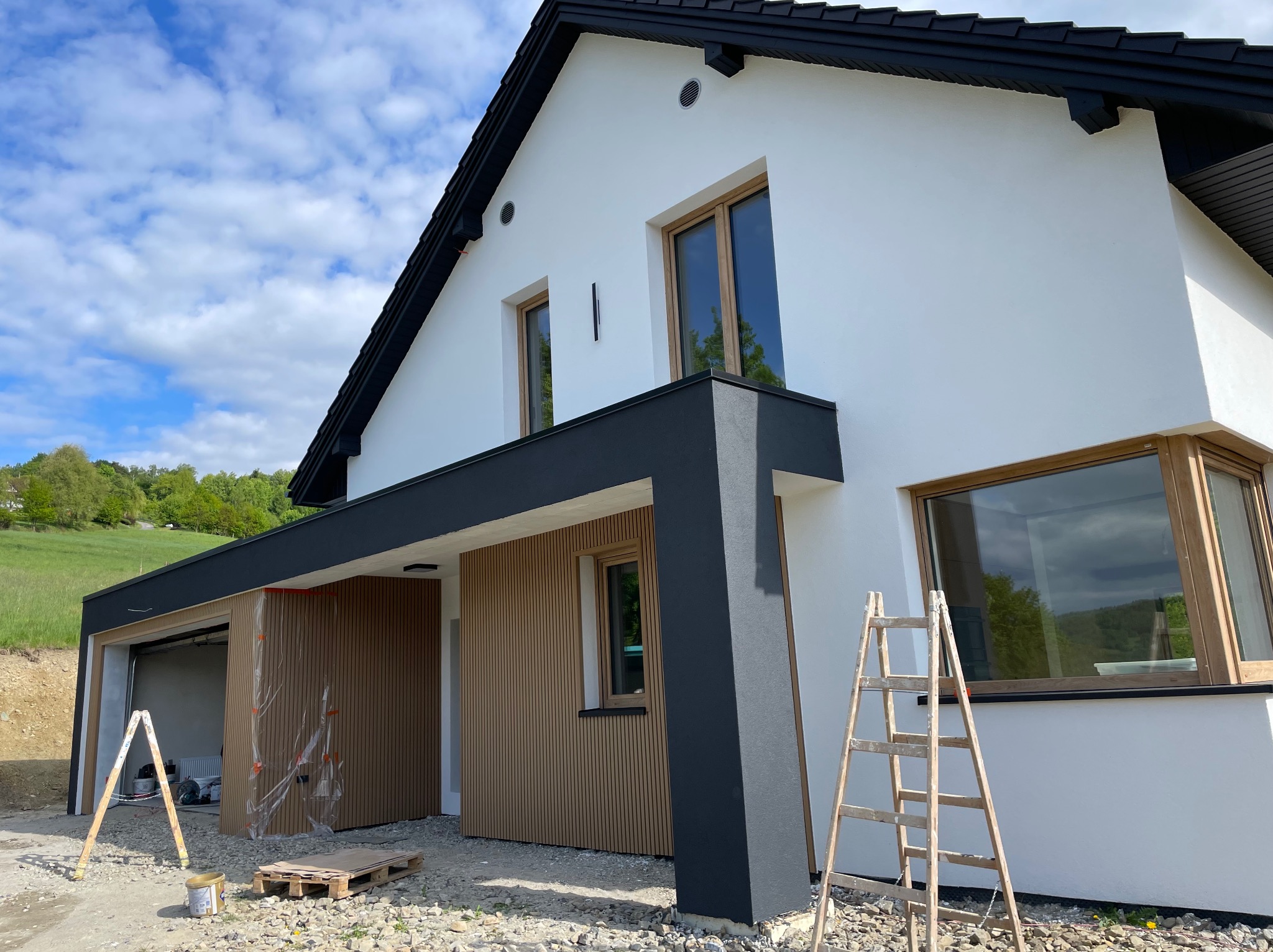 Nowoczesny dom z białą elewacją, czarnym dachem i drewnianymi akcentami. Widoczne rusztowanie i elementy wykończeniowe, sugerujące prace budowlane w toku.