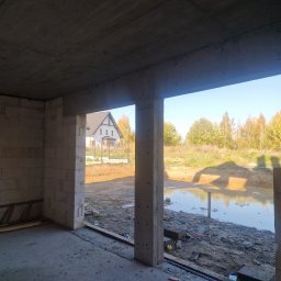 Szymon Stalke - Surowy stan domu w budowie: ściany z betonu komórkowego, widok na staw i dom w tle przez otwór okienny. Na podłodze widoczne deski i drabina.