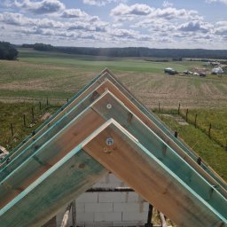 Szymon Stalke - Konstrukcja dachu z impregnowanego drewna na tle krajobrazu z polami i lasem, widok z góry, jasne niebo z chmurami. Elementy rusztowania widoczne w dolnej części kadru.