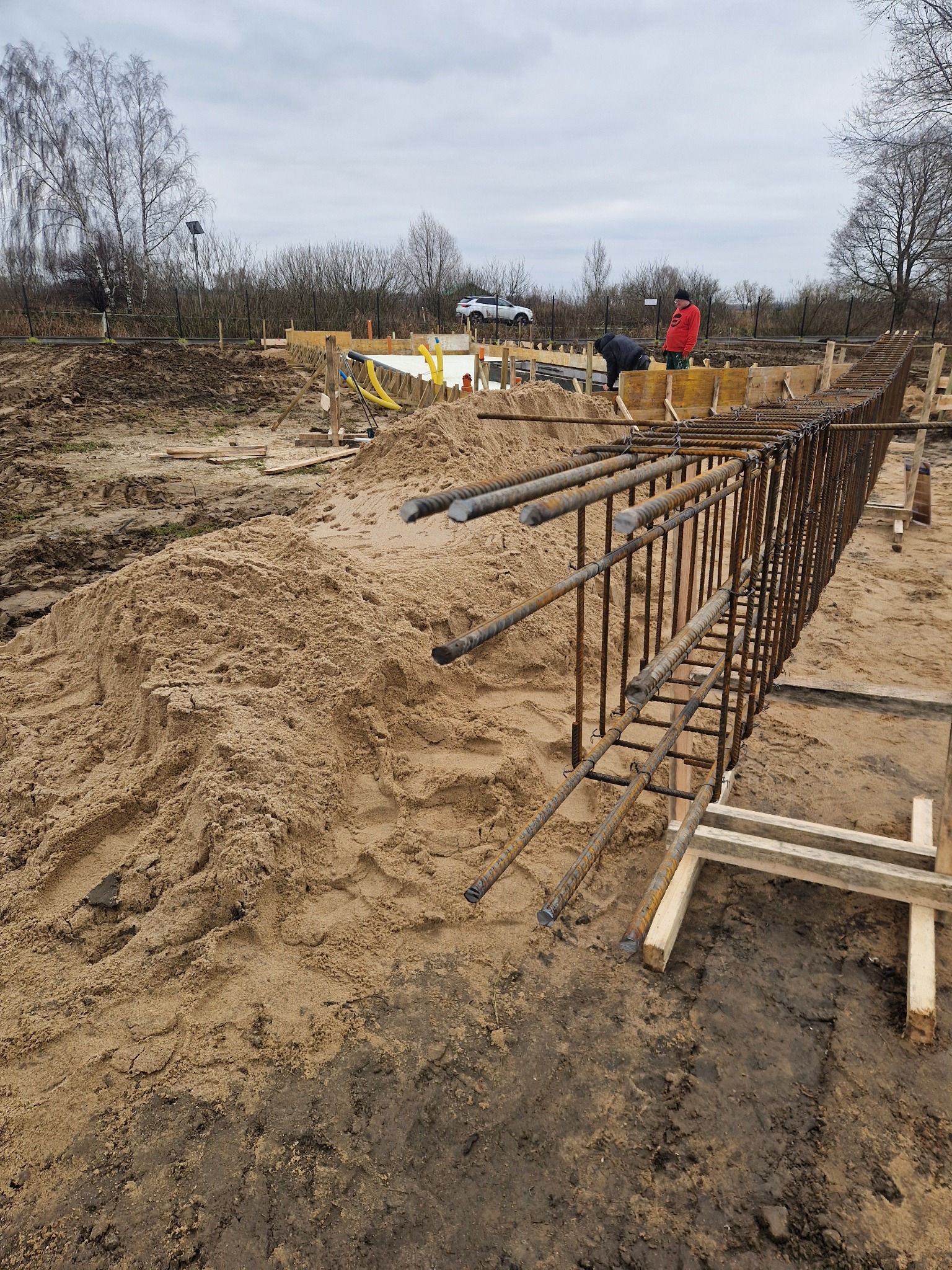 Zbrojenie fundamentów z wystającymi prętami, przygotowane do zalania betonem. Widoczny piasek, drewniane szalunki i pracownicy w tle. Pochmurne niebo.