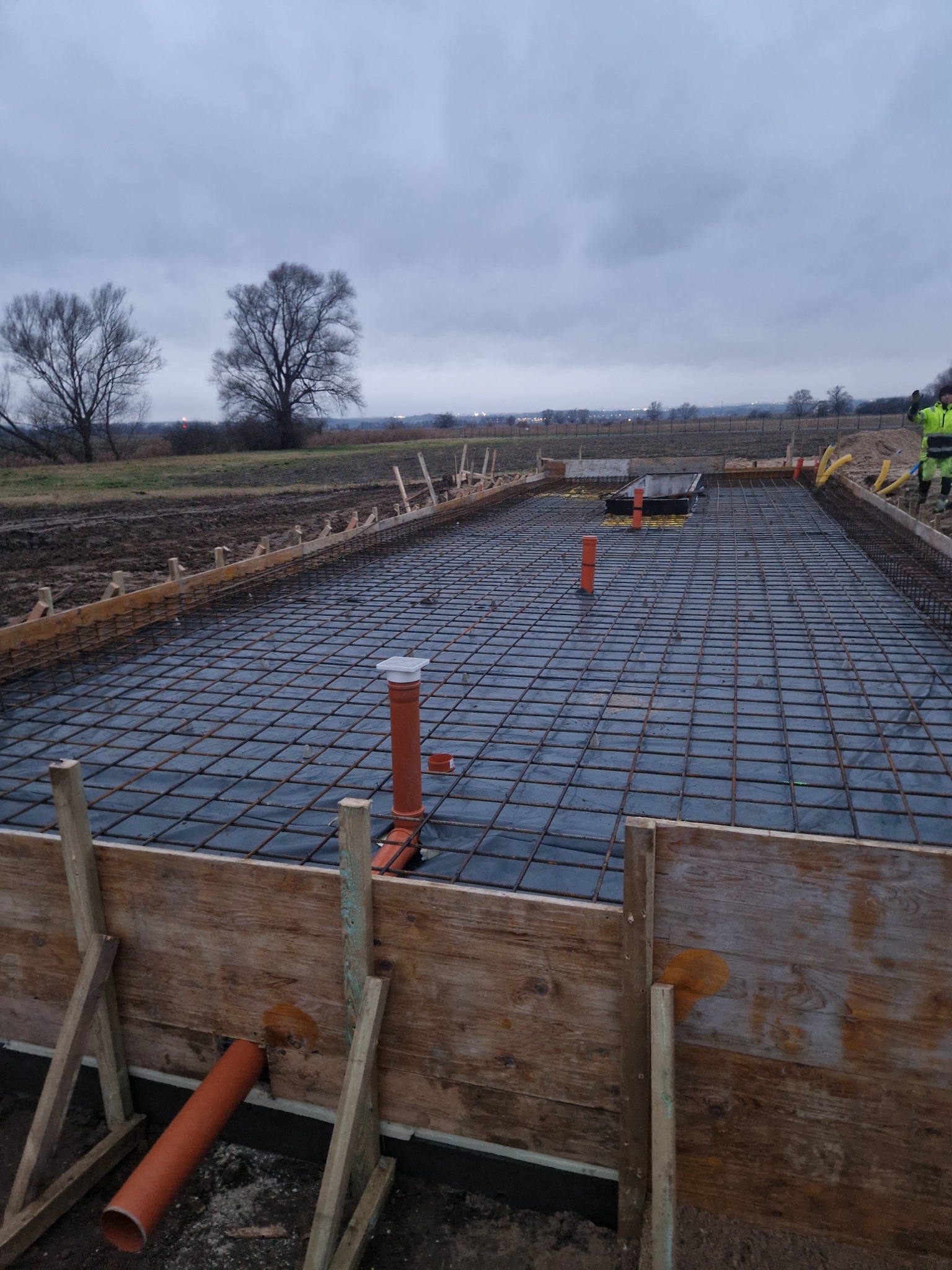 Zbrojenie fundamentów pod budowę domu jednorodzinnego. Widoczne szalunki, siatka zbrojeniowa i instalacje kanalizacyjne. Pochmurne niebo w tle.